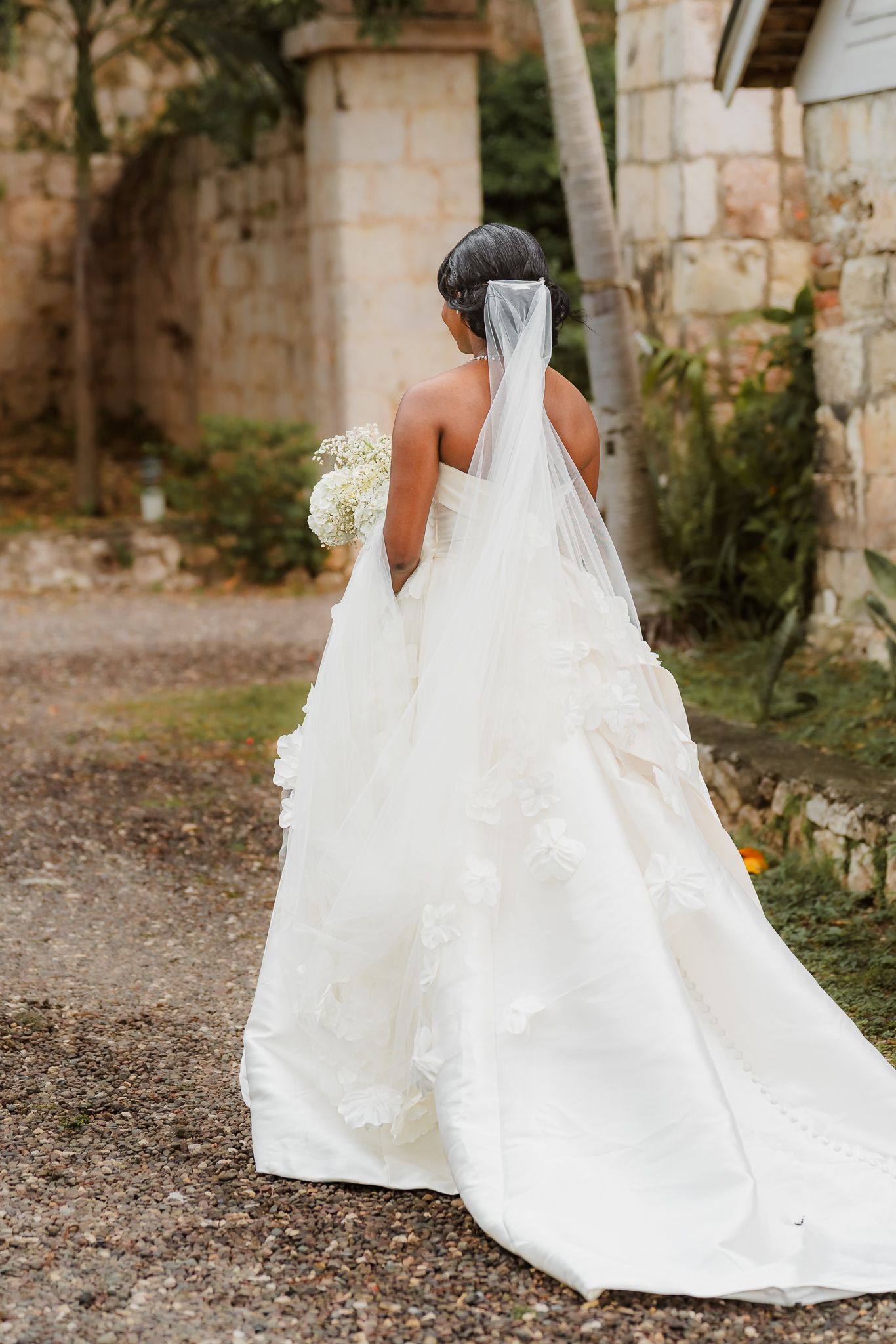 Bride in elegant white lace wedding gown with long veil holding bouquet, viewed from behind in outdoor setting.
