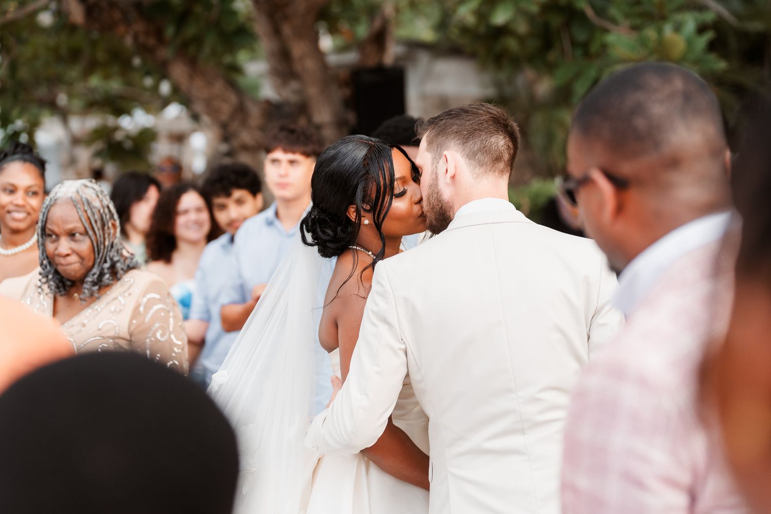 Interracial couple sharing first kiss at outdoor wedding ceremony surrounded by guests.