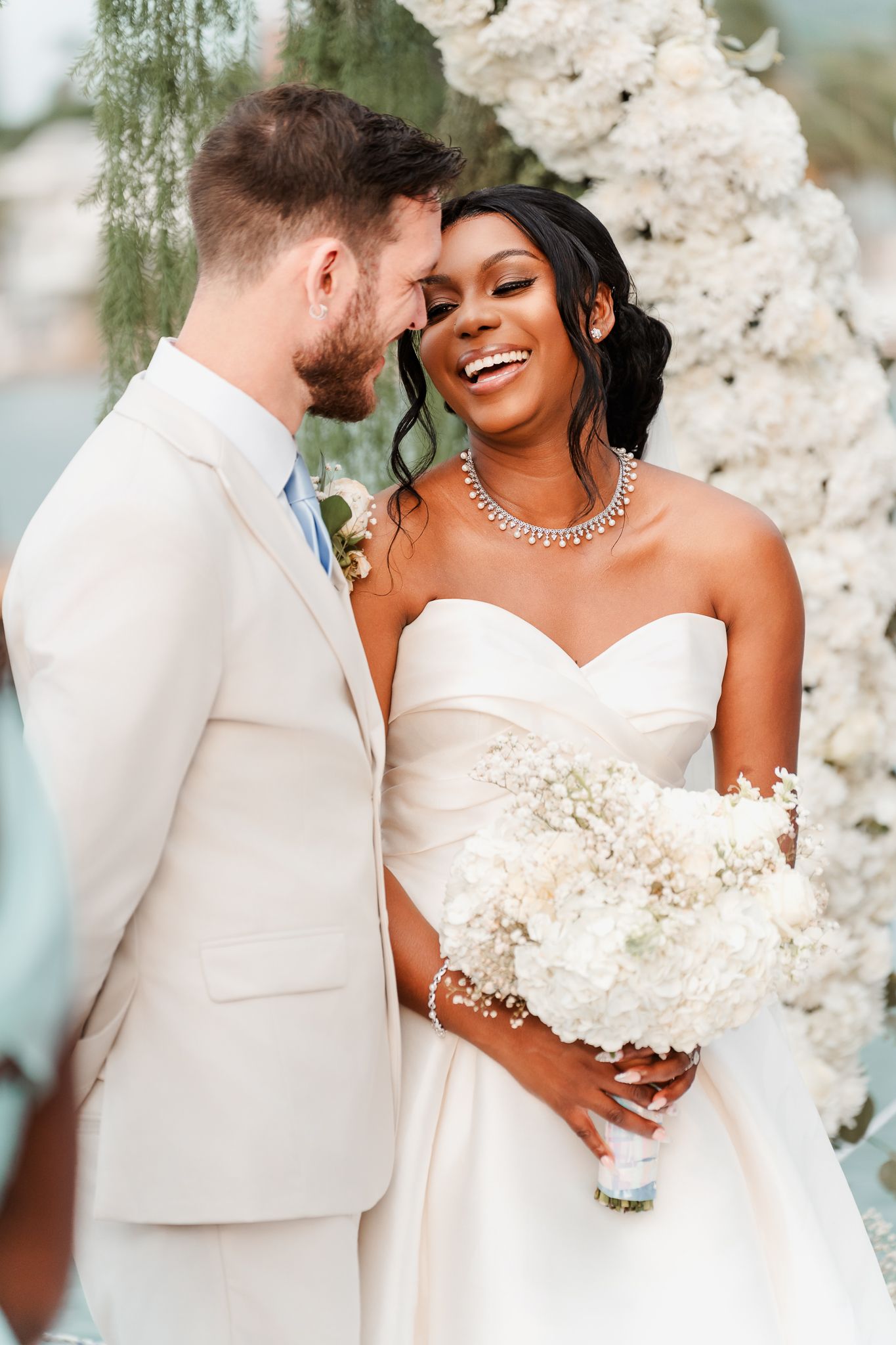 Joyful interracial couple at wedding ceremony, bride in strapless gown holding white bouquet, groom in cream suit.
