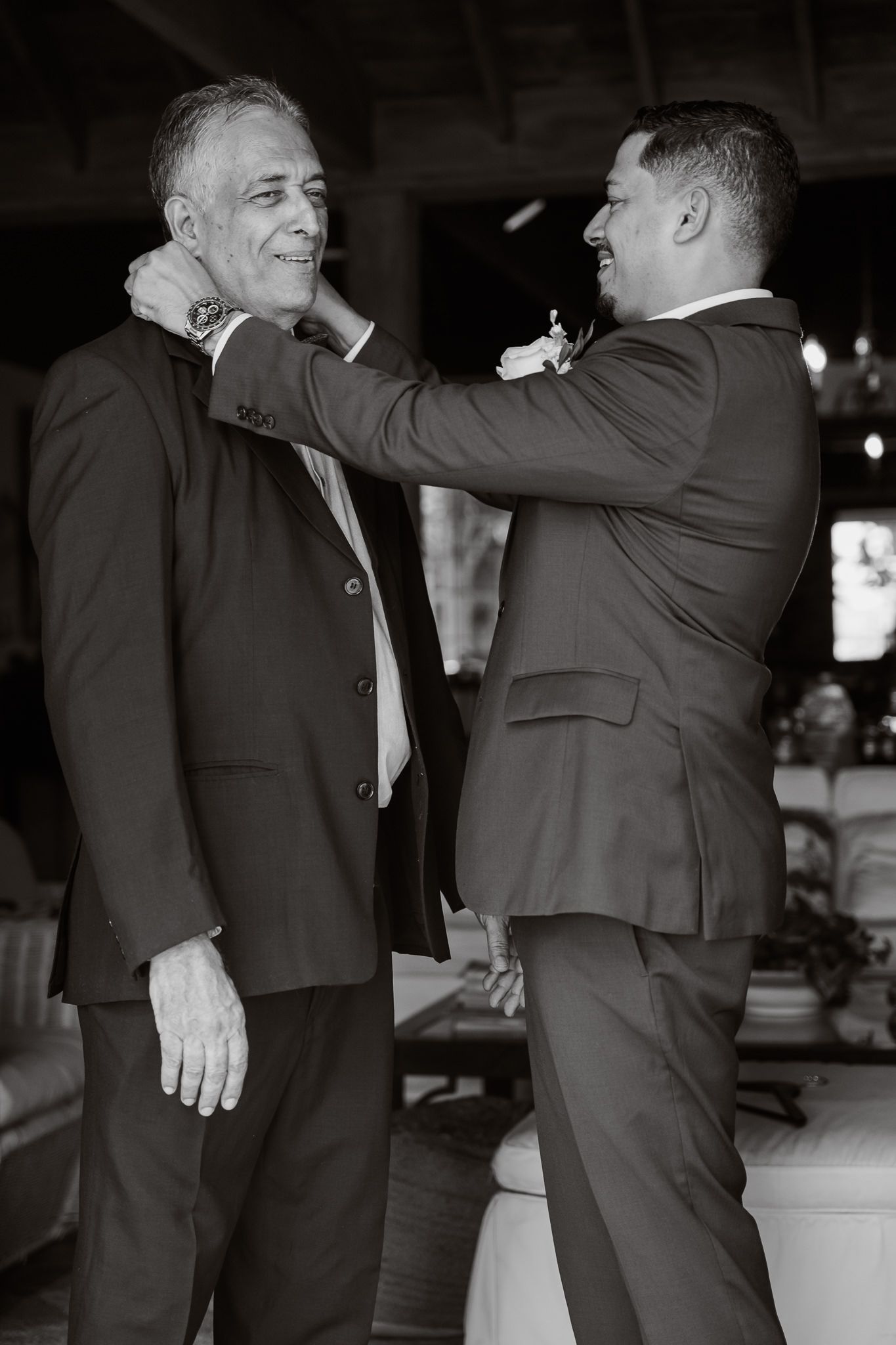 A black and white photo shows two men in suits adjusting each other's ties before a formal event.