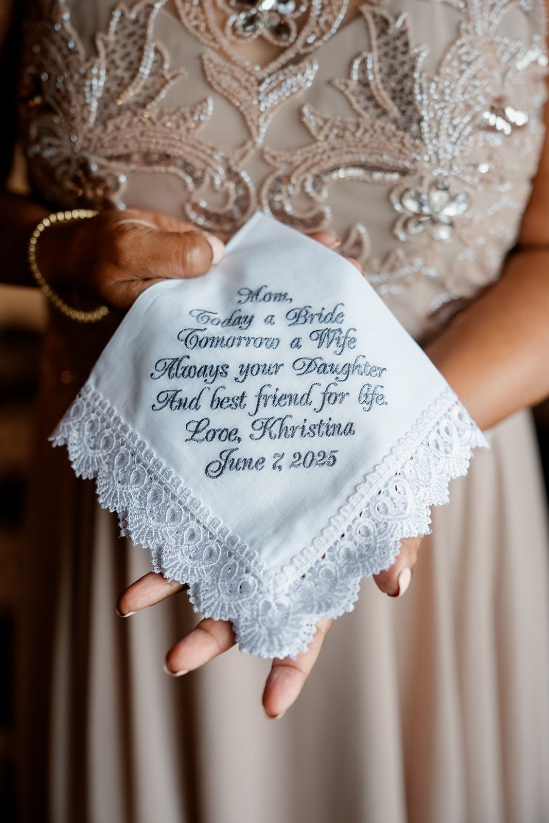 A delicate white lace handkerchief with embroidered wedding message displayed against a beige dress background.