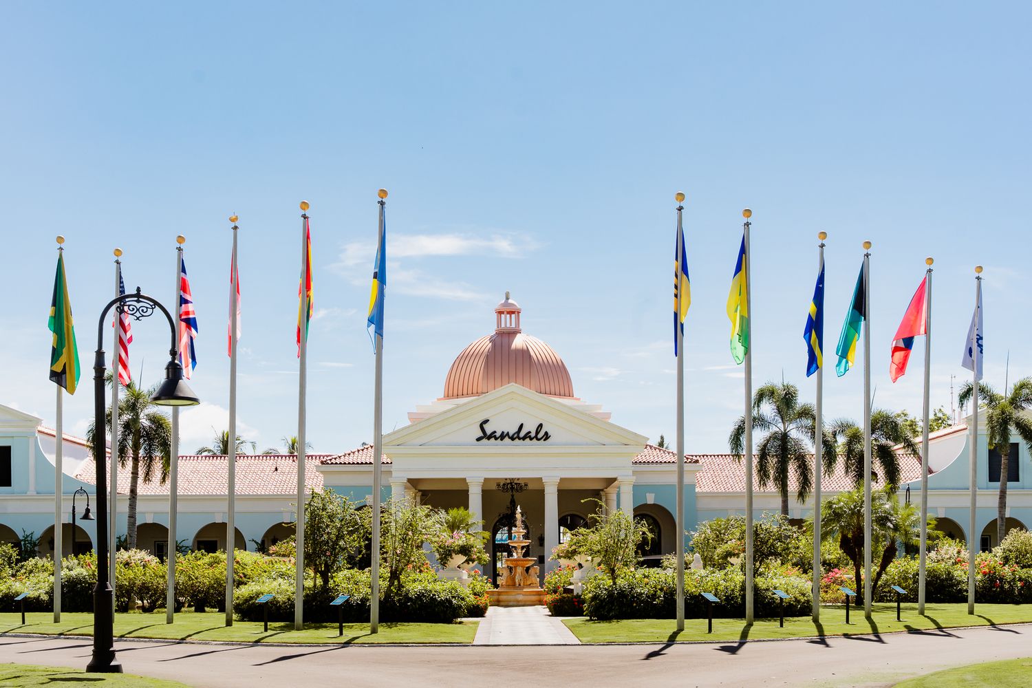 Sandals resort entrance with colorful international flags, lush palm trees, and a elegant copper-domed building under blue skies.
