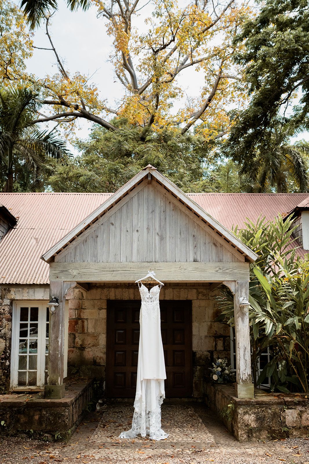 A white wedding dress hangs from a rustic stone cottage with a tin roof surrounded by tropical palm trees and foliage.