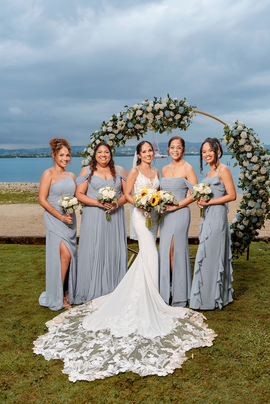 Bridal party poses under floral arch with ocean view backdrop, bridesmaids wearing light gray gowns with bride in white lace gown.