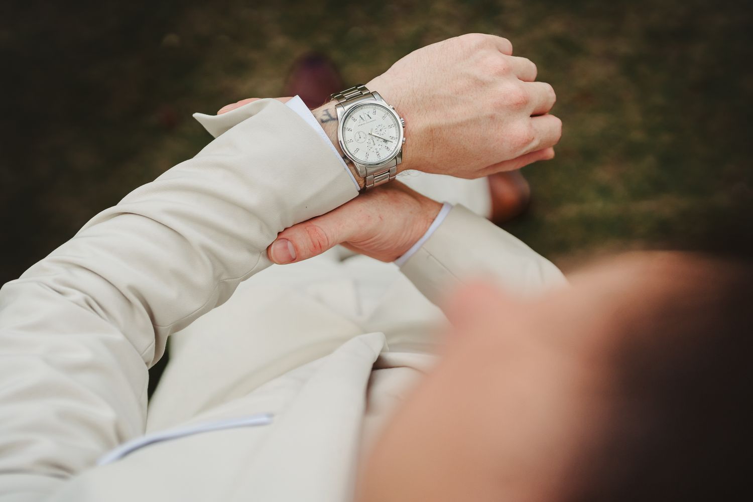 Man in white suit checking silver wristwatch outdoors on green grass background.