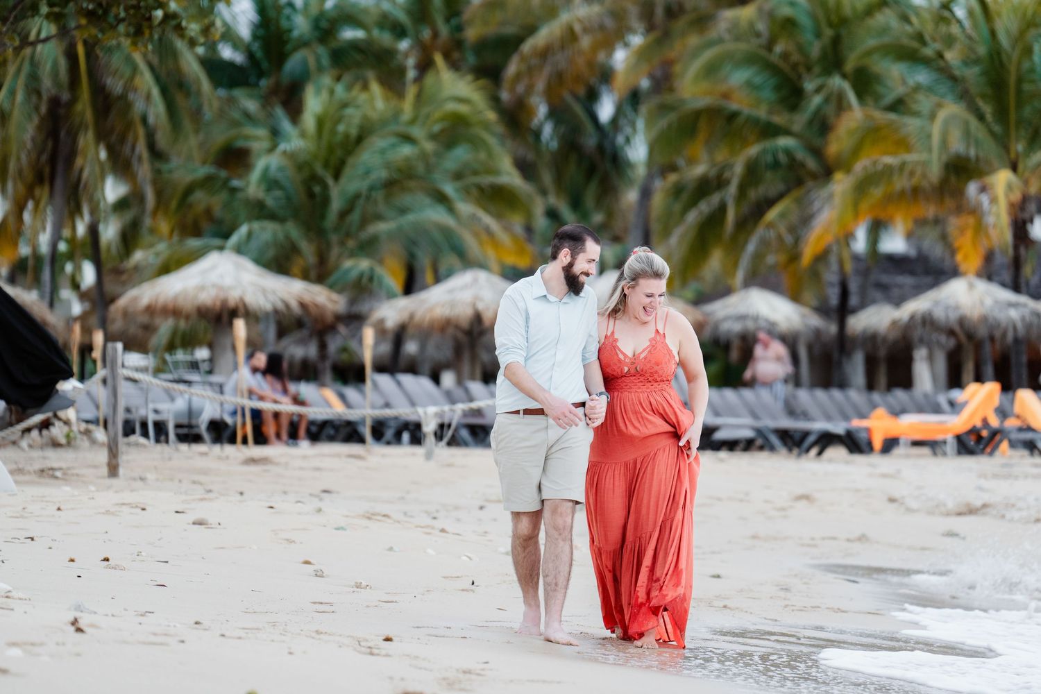 A couple walks along a tropical beach with palm trees and thatched umbrellas in the background.