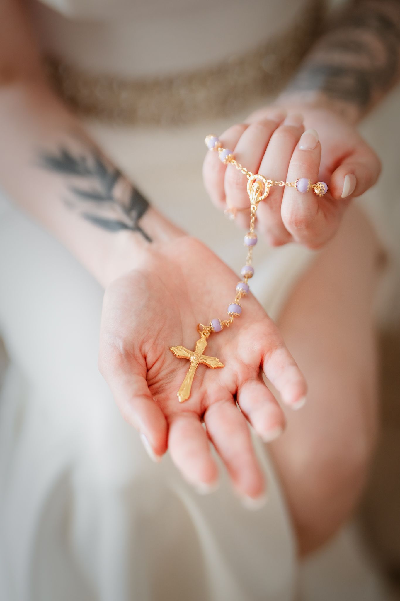 Tattooed bride holding a gold cross rosary with pastel beads in her open palms, wearing a white wedding dress.