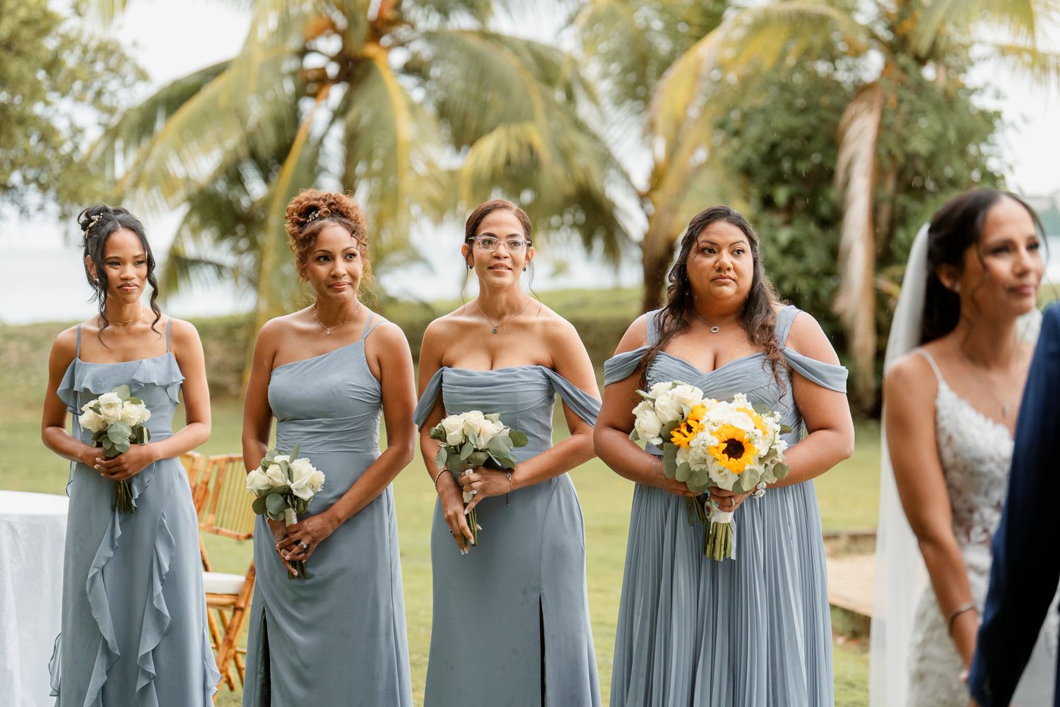Row of bridesmaids in dusty blue dresses holding bouquets standing outdoors with palm trees in background.