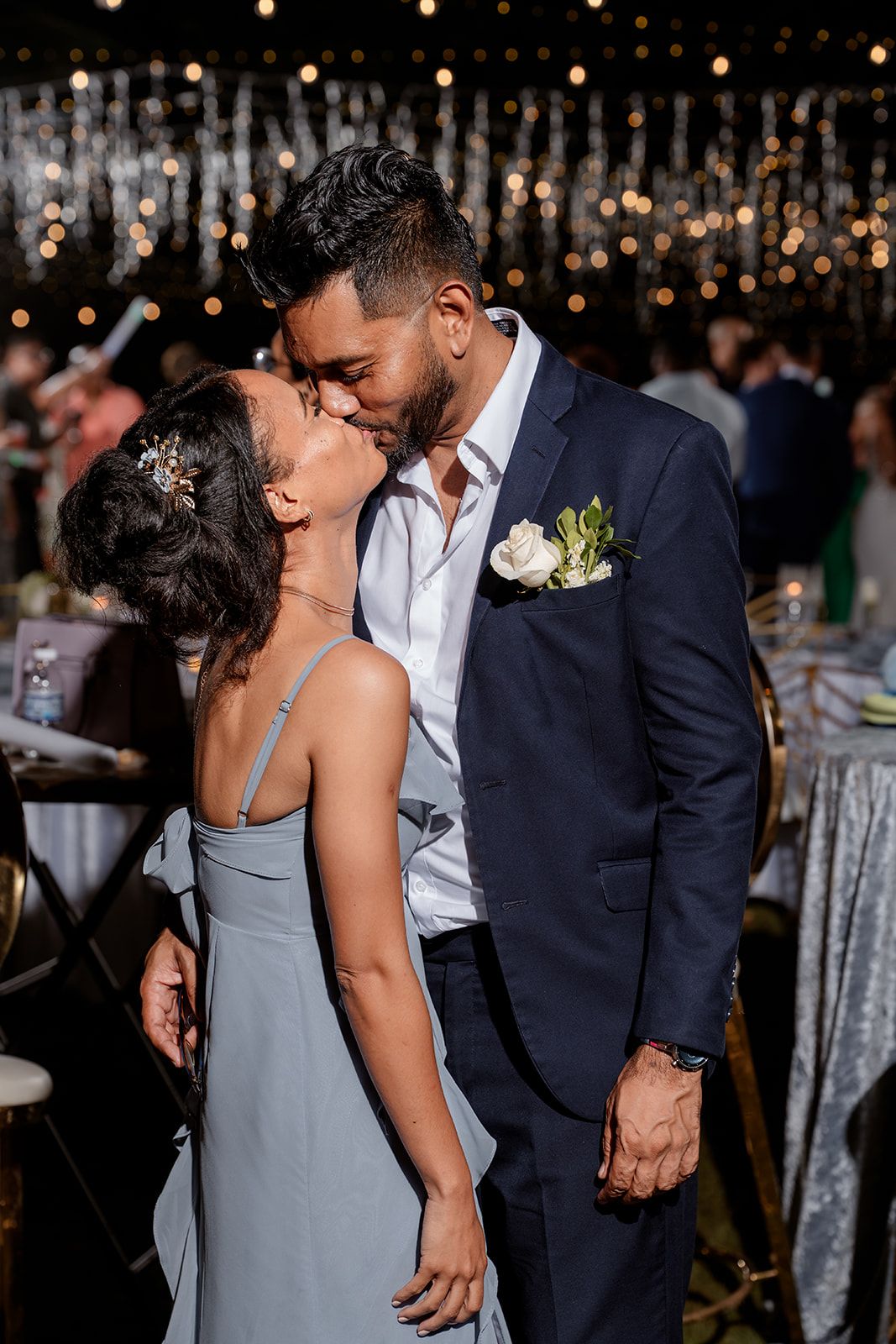 A couple shares a romantic kiss at an elegant wedding reception with sparkling lights in the background.