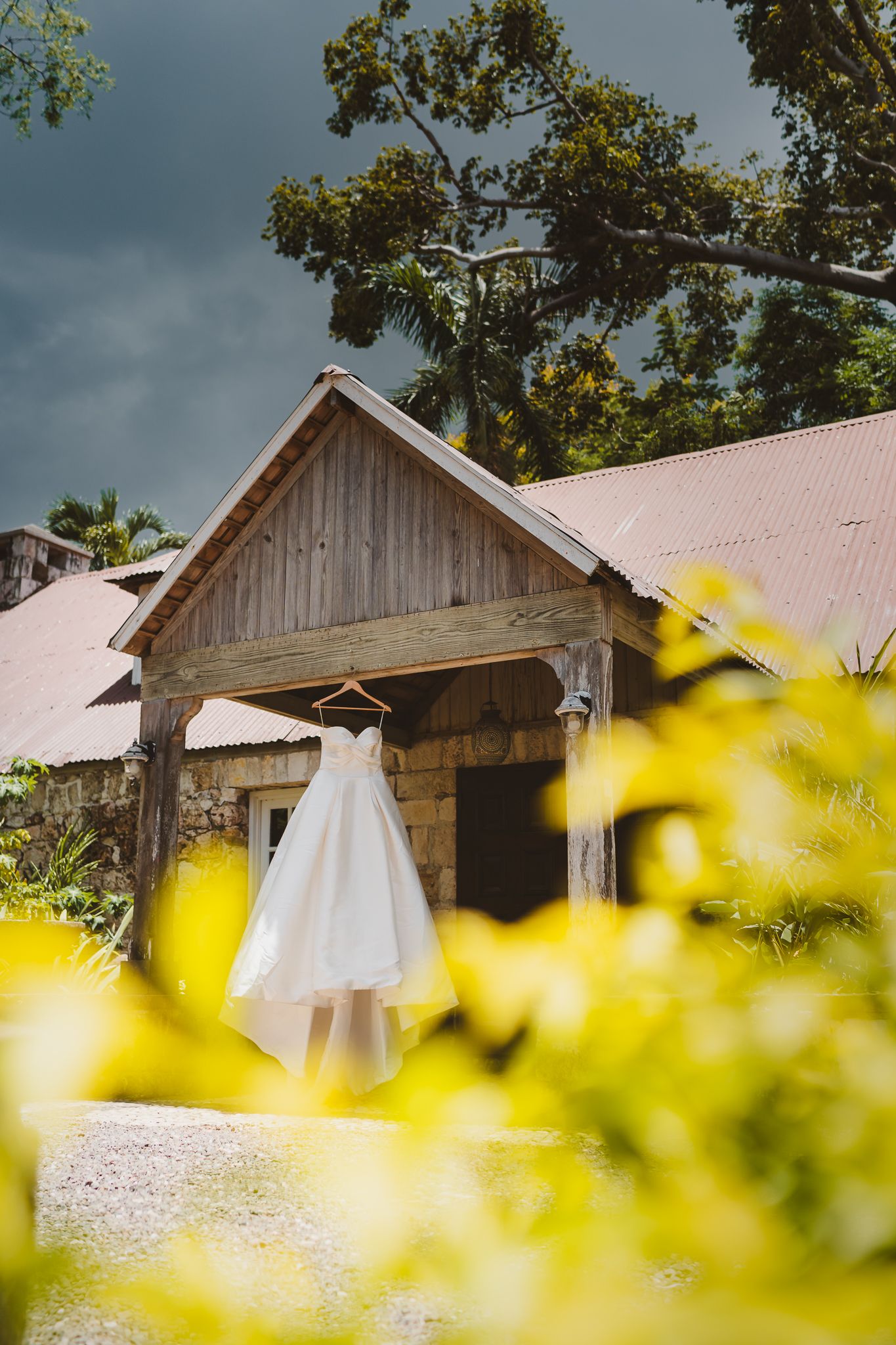 White wedding dress hanging on rustic wooden barn entrance surrounded by lush tropical greenery under dramatic stormy sky.