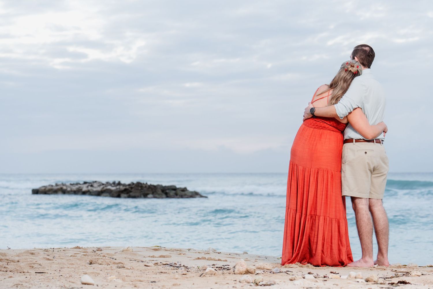 A couple embraces while standing on a tropical beach with coral colored dress flowing in the breeze near rocky coastline.