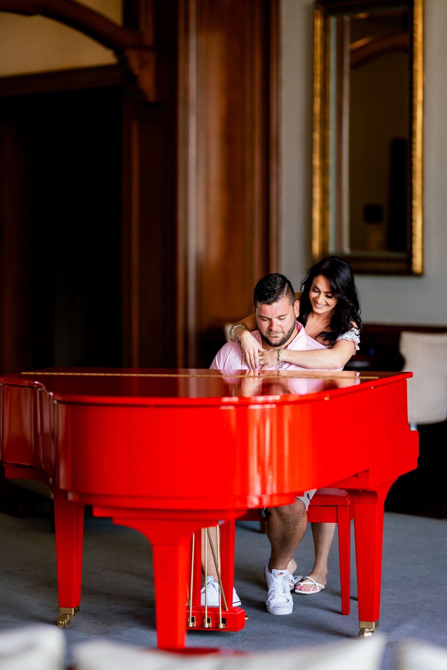 A couple shares an intimate moment at a bright red grand piano in an elegant interior setting.