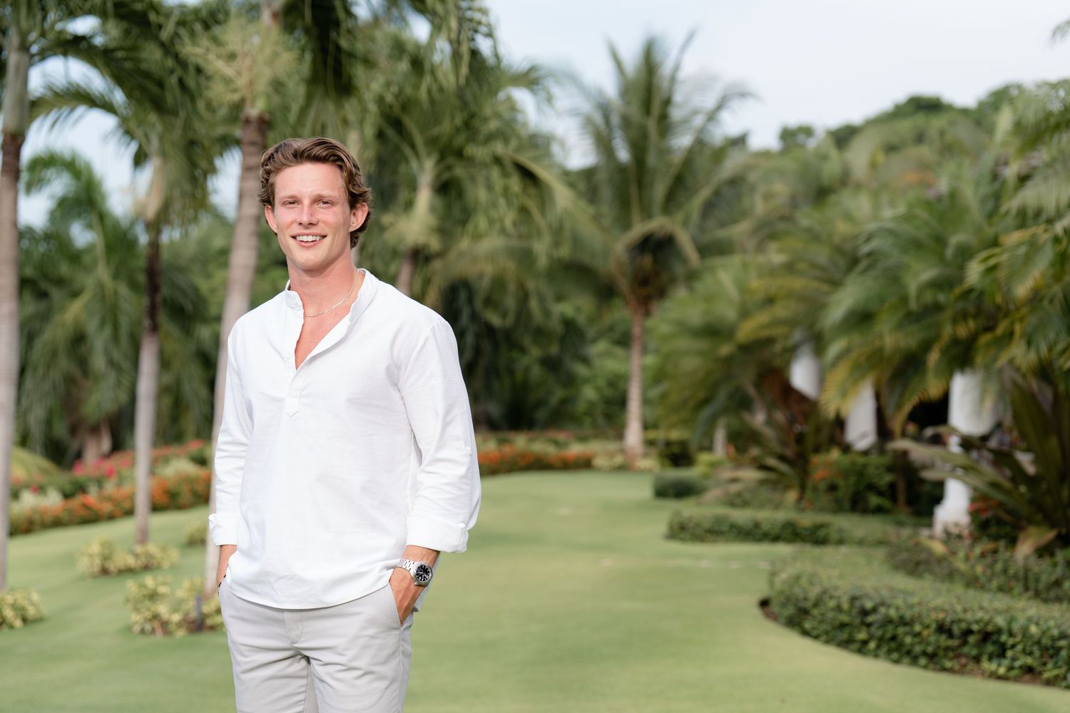 Person in white button-down shirt and grey pants stands smiling on tropical resort lawn with palm trees in background.