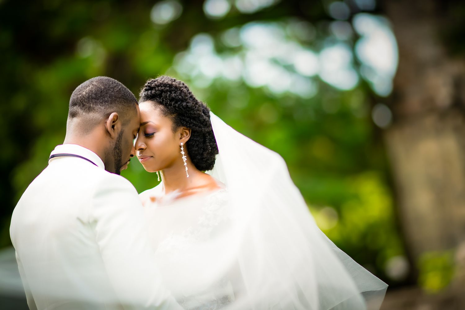 A Black bride and groom share a romantic moment outdoors, her white veil flowing gracefully in a lush green setting.