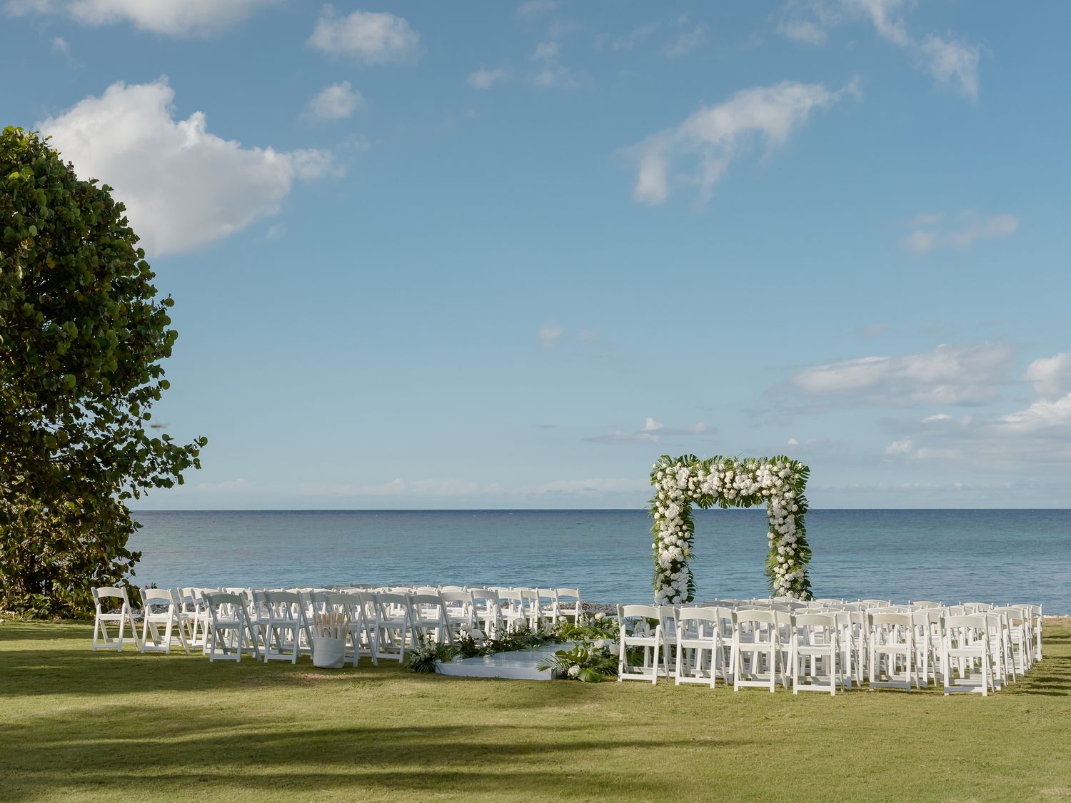 Outdoor wedding ceremony setup with white chairs, floral arch, and ocean backdrop on a lush green lawn.
