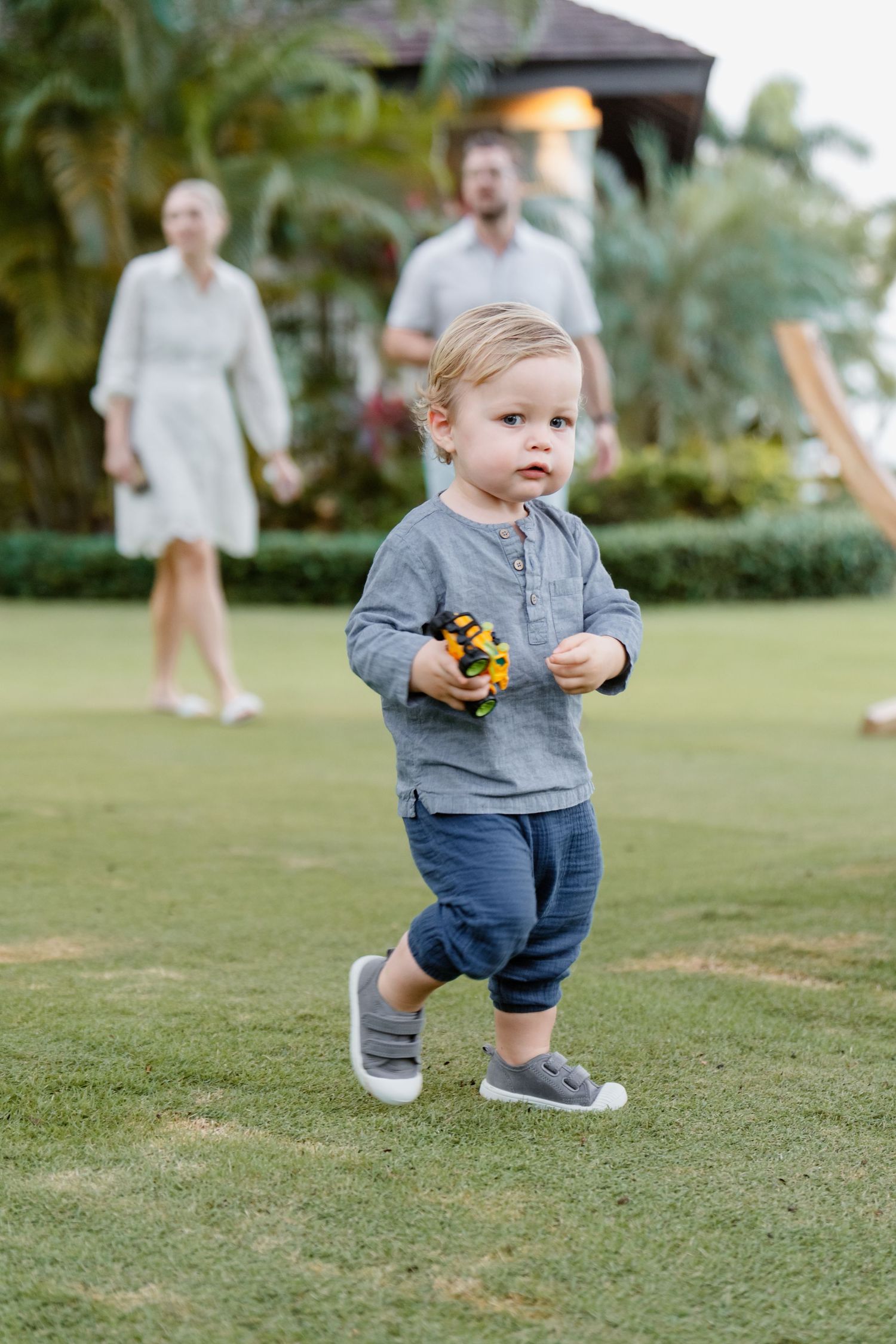 A toddler in gray shirt and jeans walks on grass while holding a toy, with two adults visible in background.