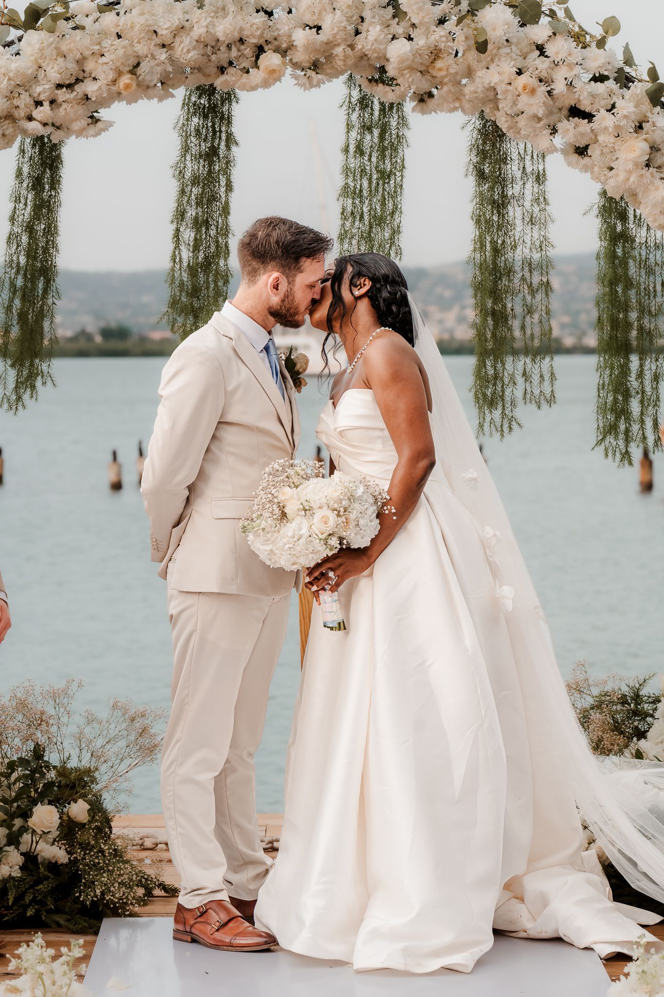Bride and groom share first kiss at waterfront wedding ceremony under floral arch with white blooms and greenery.