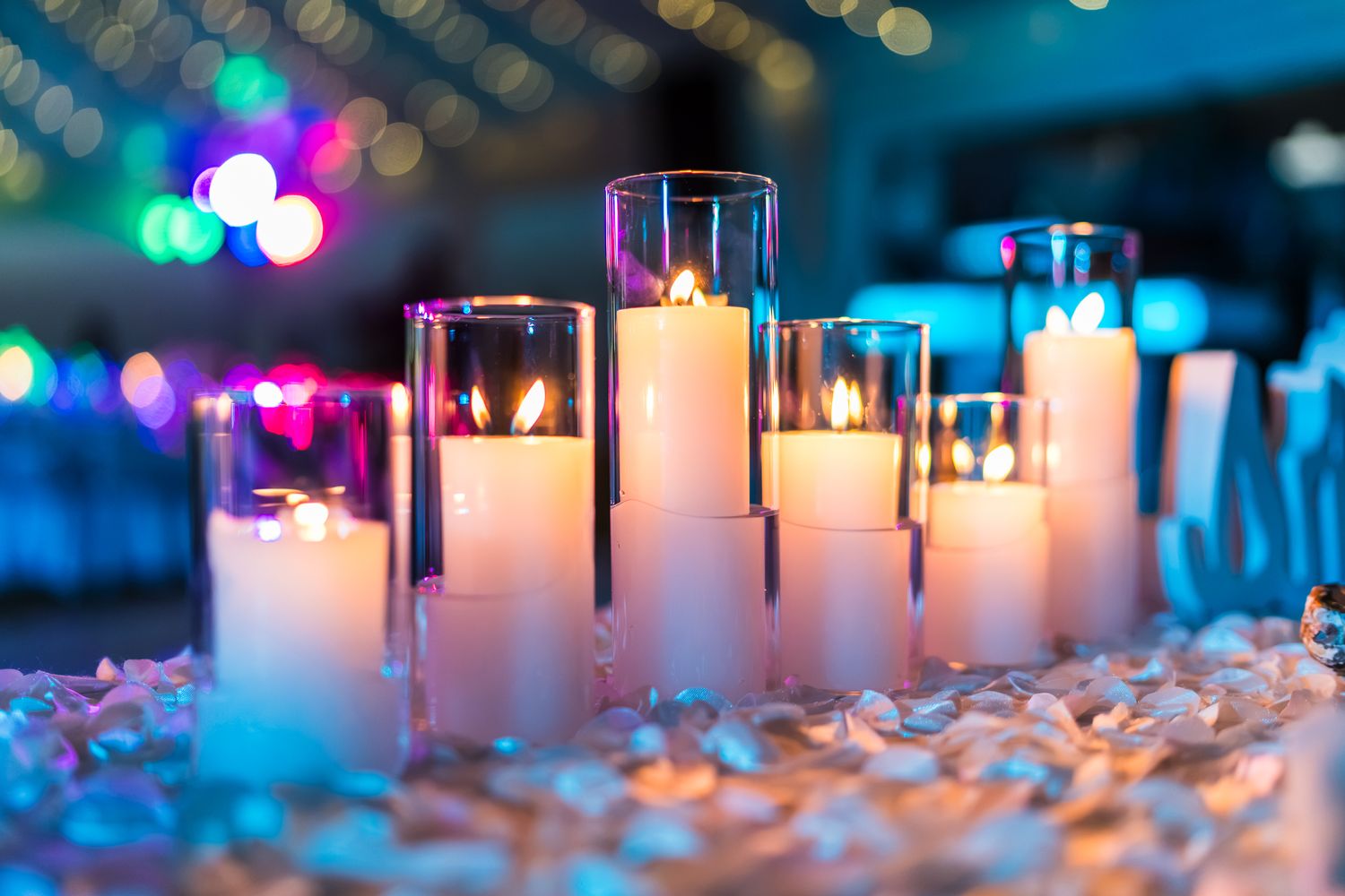 Elegant white pillar candles in glass holders glow warmly among flower petals at a colorful event.