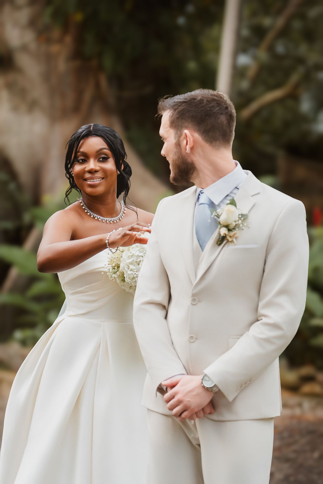 Interracial couple in wedding attire share a joyful moment outdoors, bride in white gown holding bouquet, groom in cream suit.