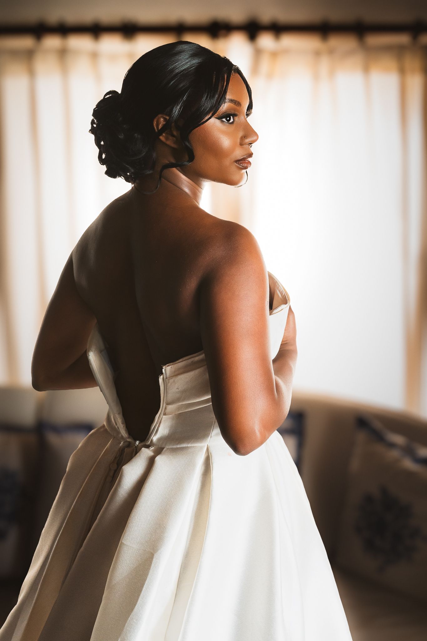 Elegant bride in strapless white satin gown with bow, looking over shoulder in soft window light.
