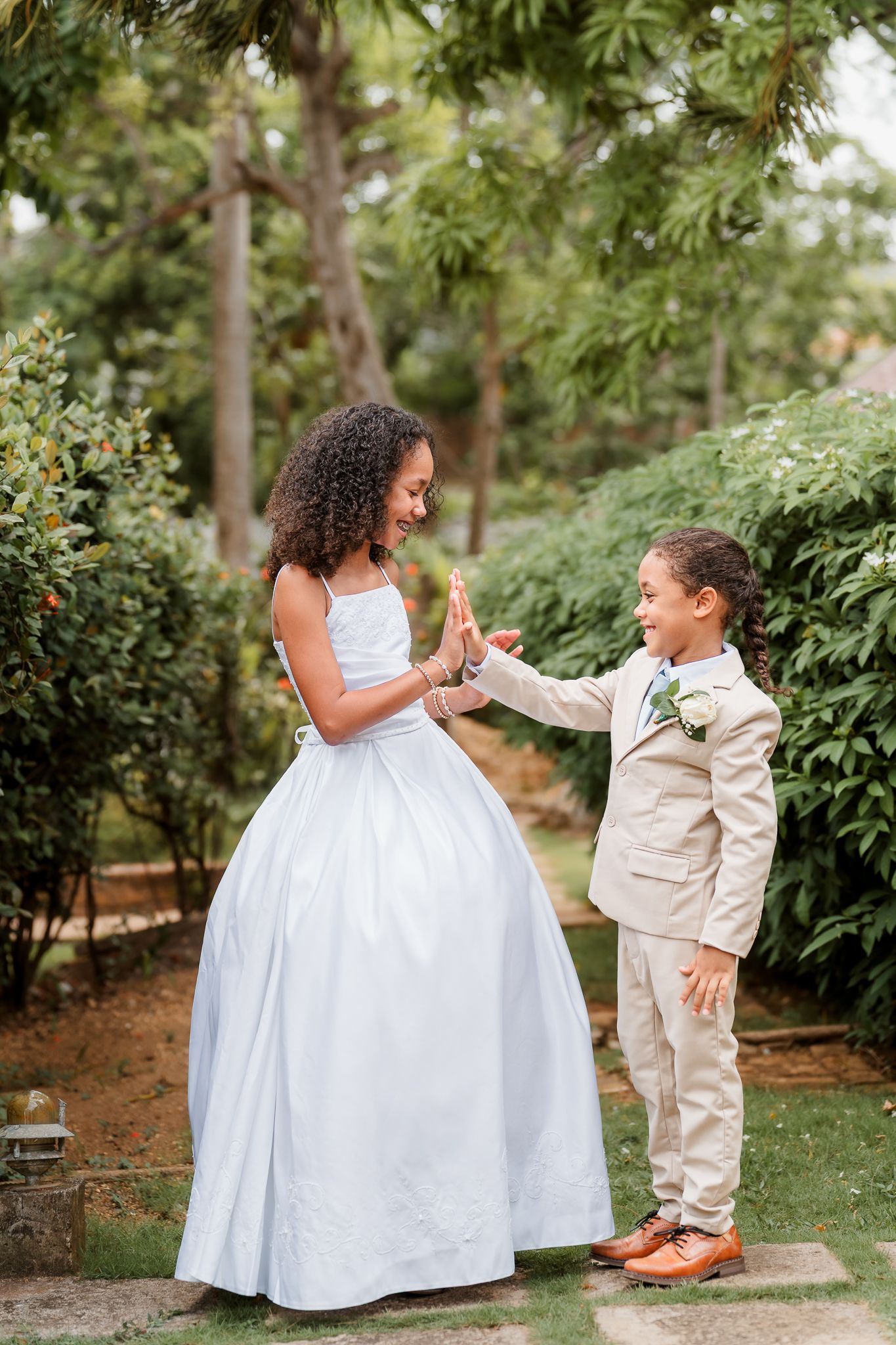 Two children in formal attire hold hands in a garden, girl in white gown and boy in beige suit with boutonniere.