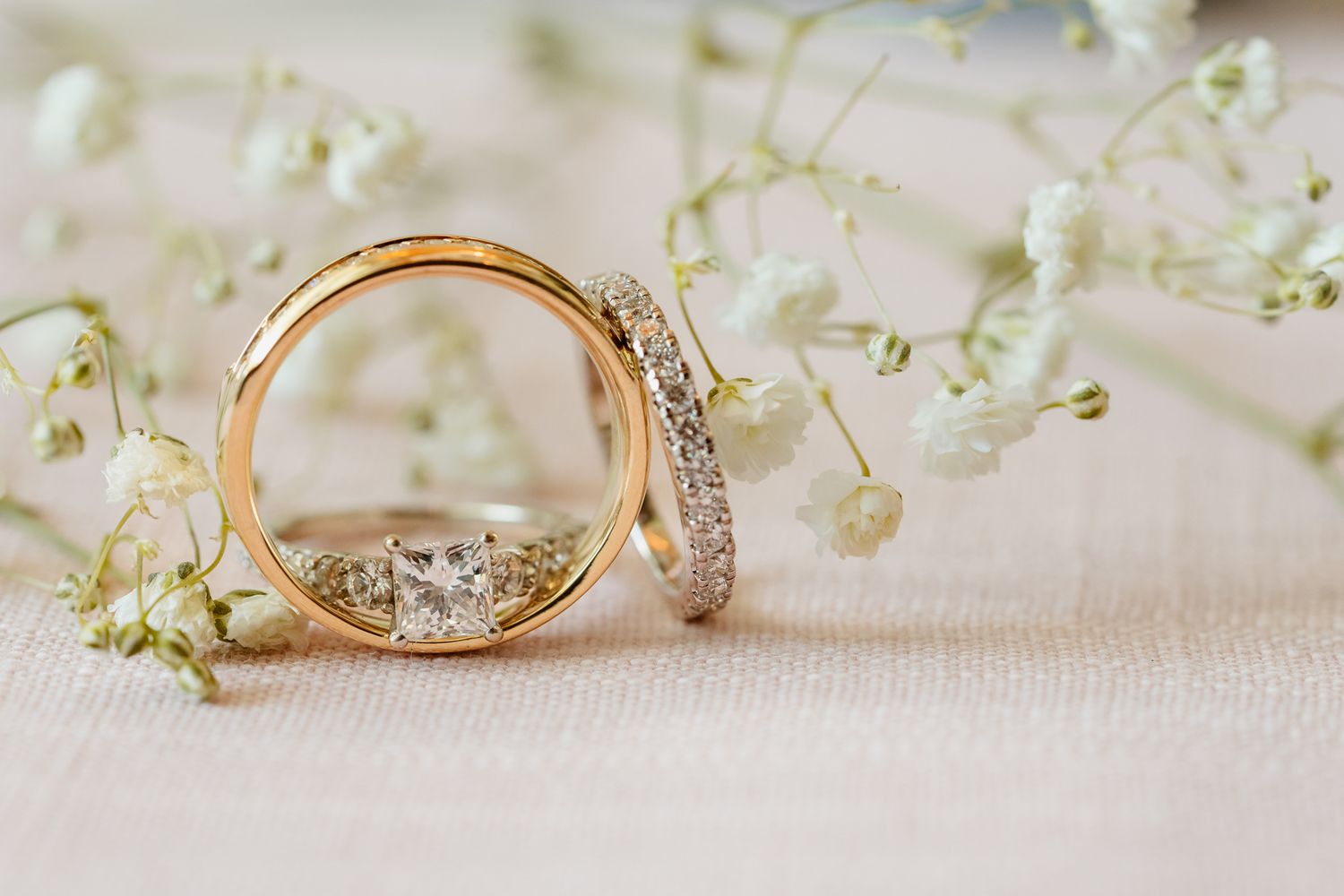Two gold wedding rings with diamonds rest against delicate white baby's breath flowers on a soft white surface.