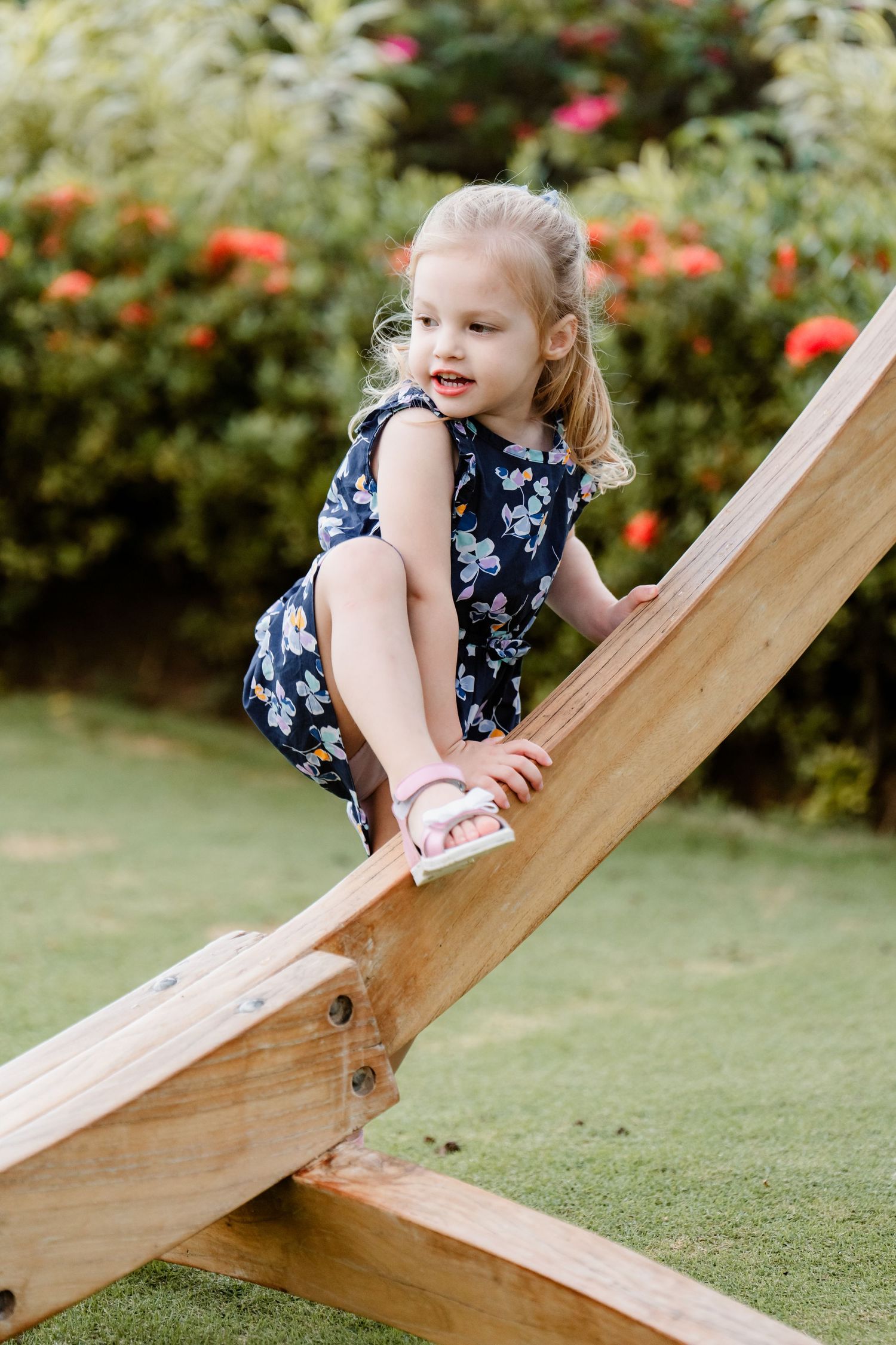 Child in floral dress climbs on wooden plank outdoors with colorful flowers and greenery in background.