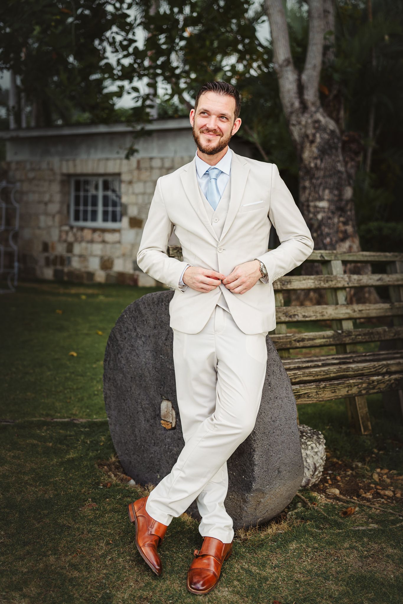 Man in beige suit with blue tie and brown monk strap shoes leaning on stone in garden setting.