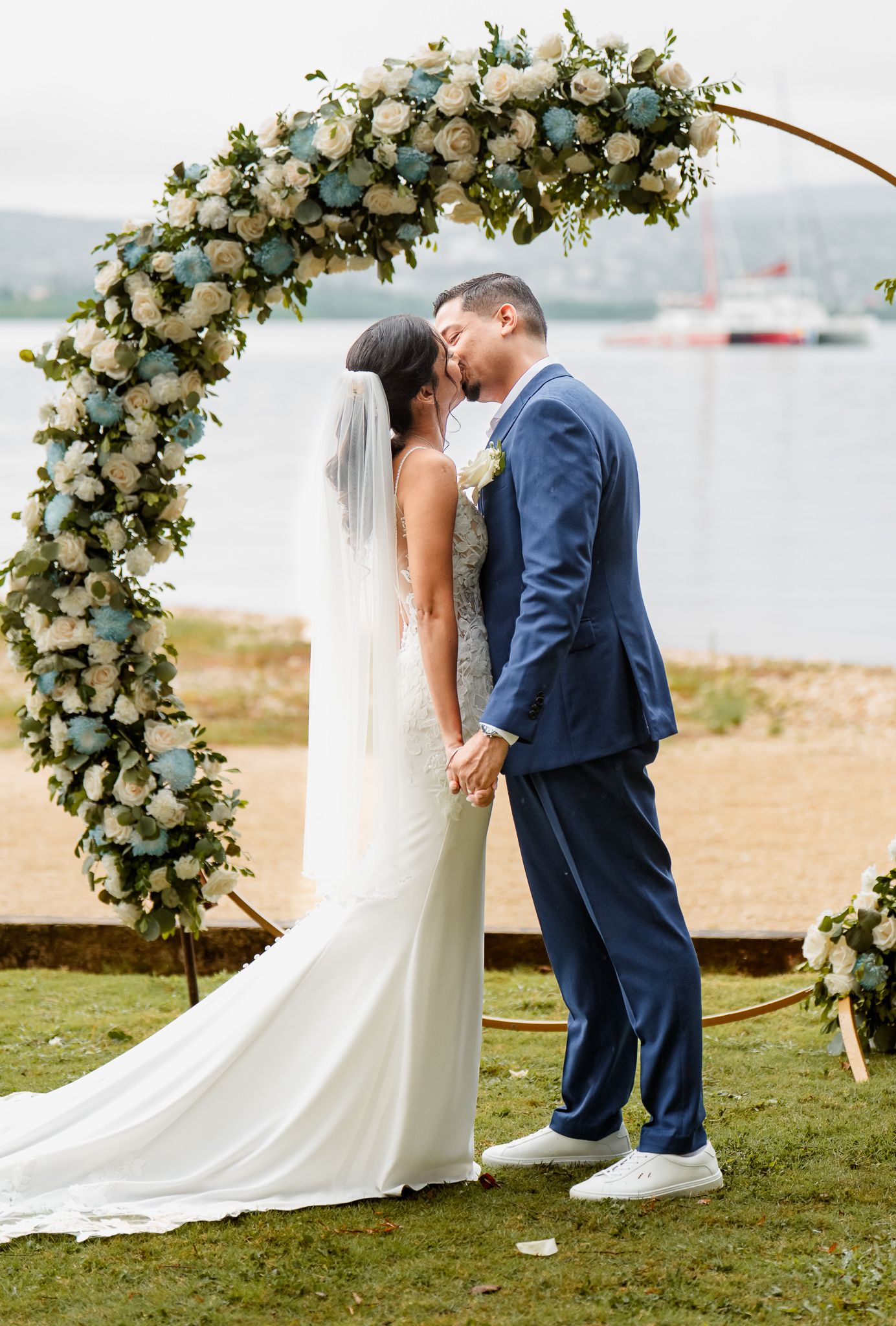 A wedding ceremony under a floral arch decorated with white roses and eucalyptus by a waterfront with boats in background.