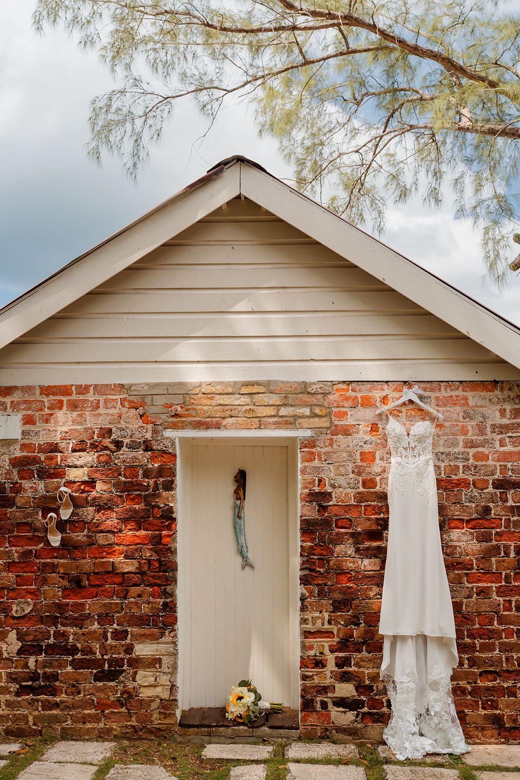 A white lace wedding dress hangs beside a simple white door on a rustic brick building with weathered siding.
