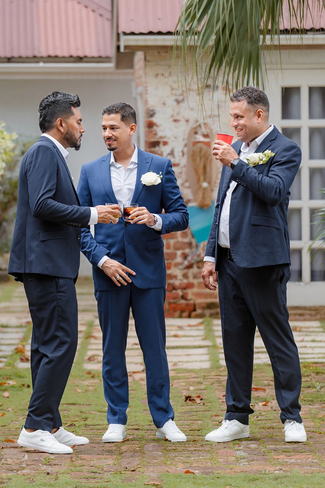 Three well-dressed men in navy and blue suits with white sneakers share drinks and conversation at an outdoor event.