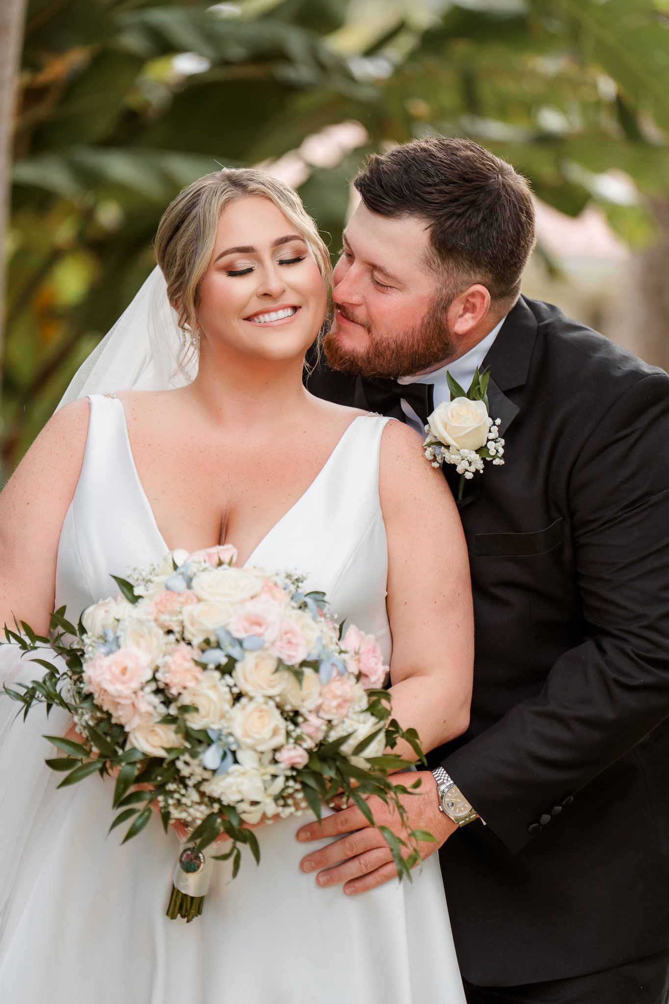 Bride and groom share a romantic moment, groom kissing bride's cheek as she holds a pastel floral bouquet.