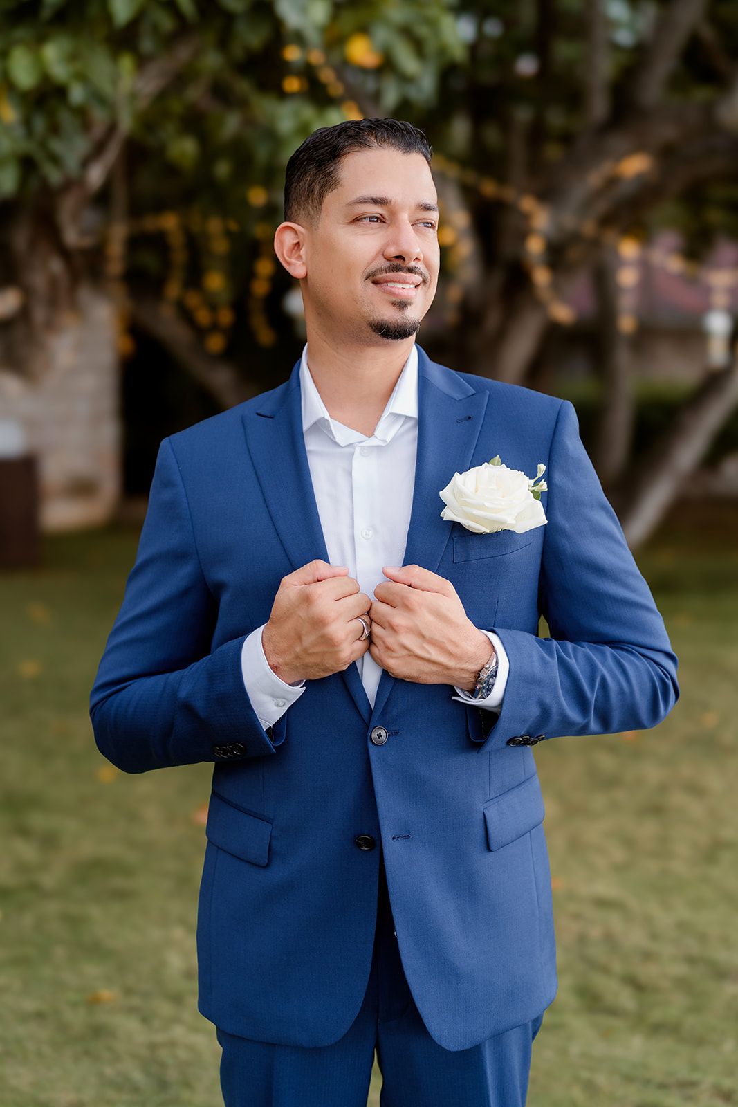 Groom in a tailored royal blue suit and white boutonniere adjusts his jacket while standing outdoors.