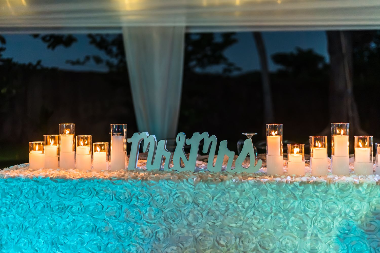 Romantic wedding sweetheart table with glowing candles and Mr. & Mrs. sign on teal rosette tablecloth.