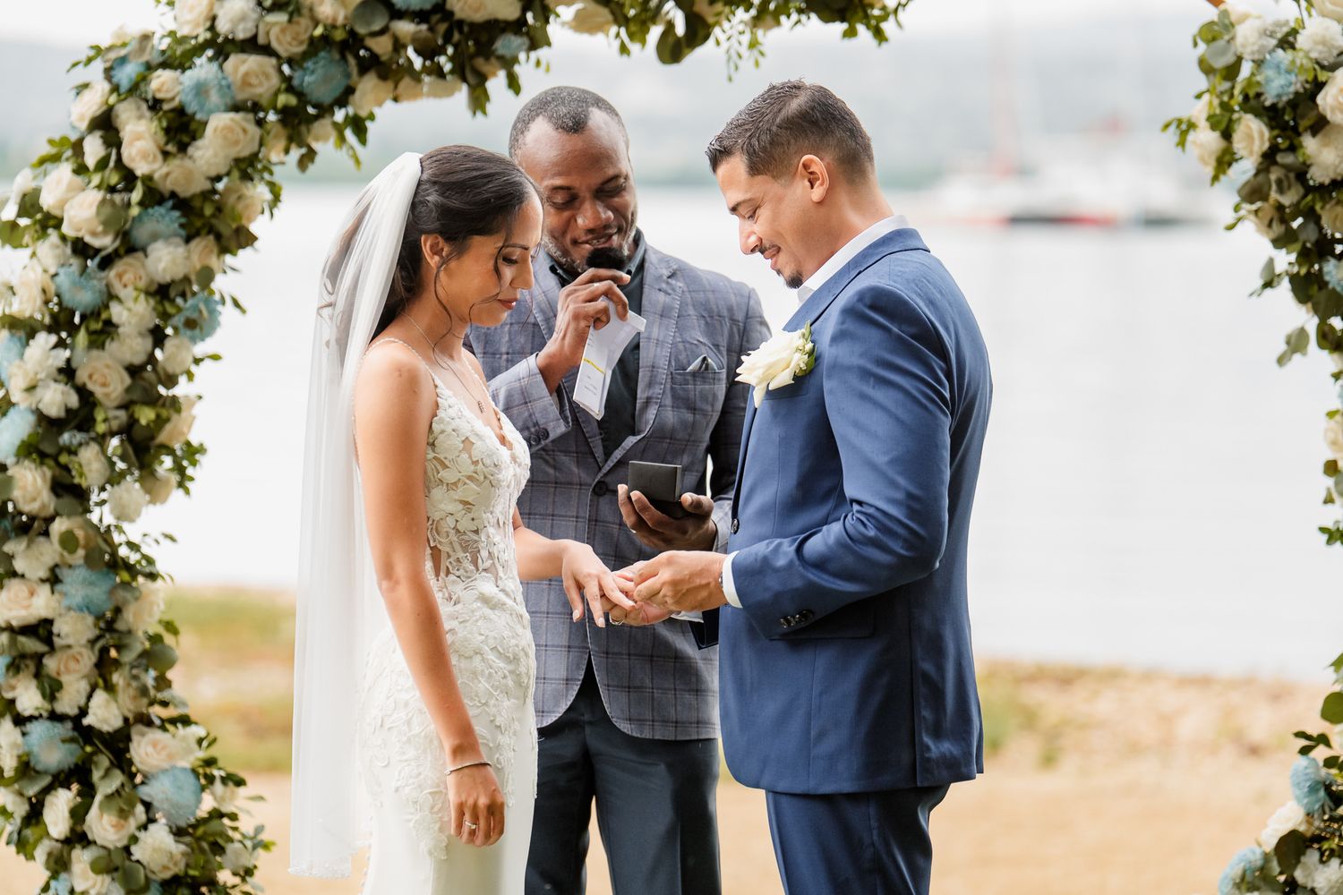 Wedding ceremony taking place under a floral arch with white roses and greenery on a bright sunny day.