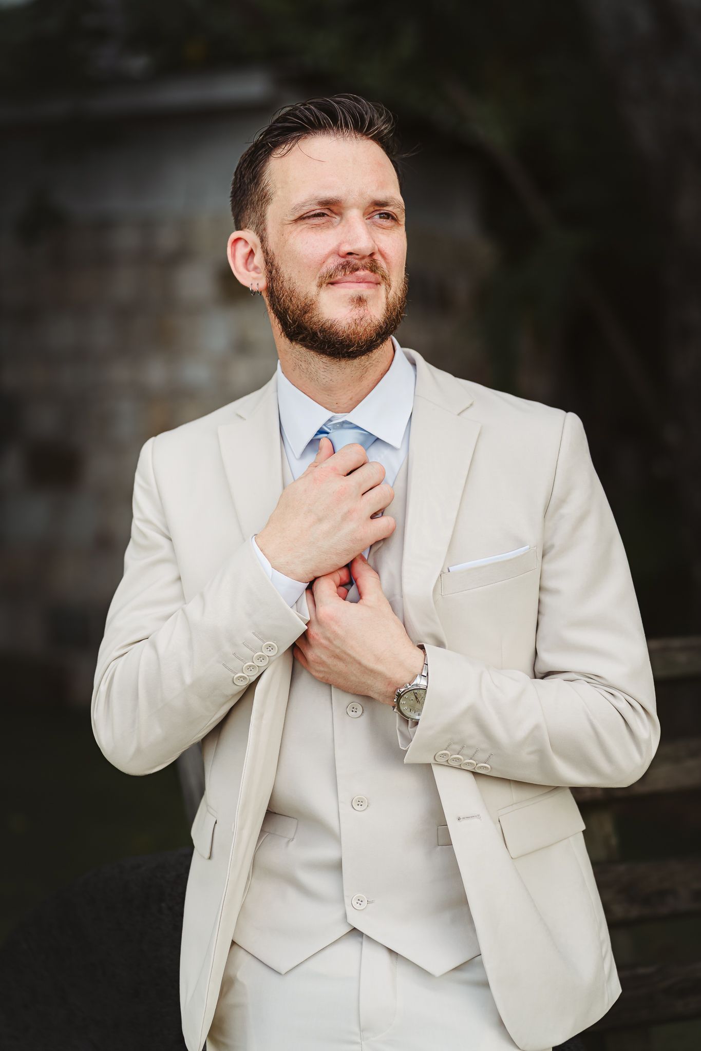 A bearded groom in a beige three-piece suit adjusts his blue tie while looking up outdoors.