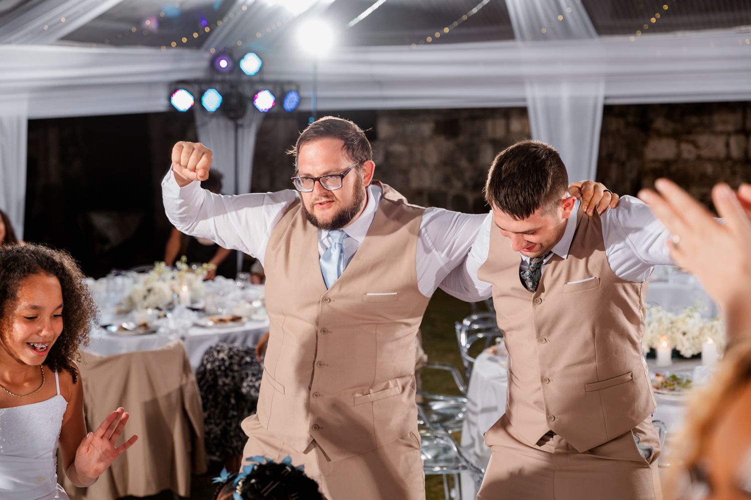 Two groomsmen in tan vests dancing joyfully at a wedding reception celebration.