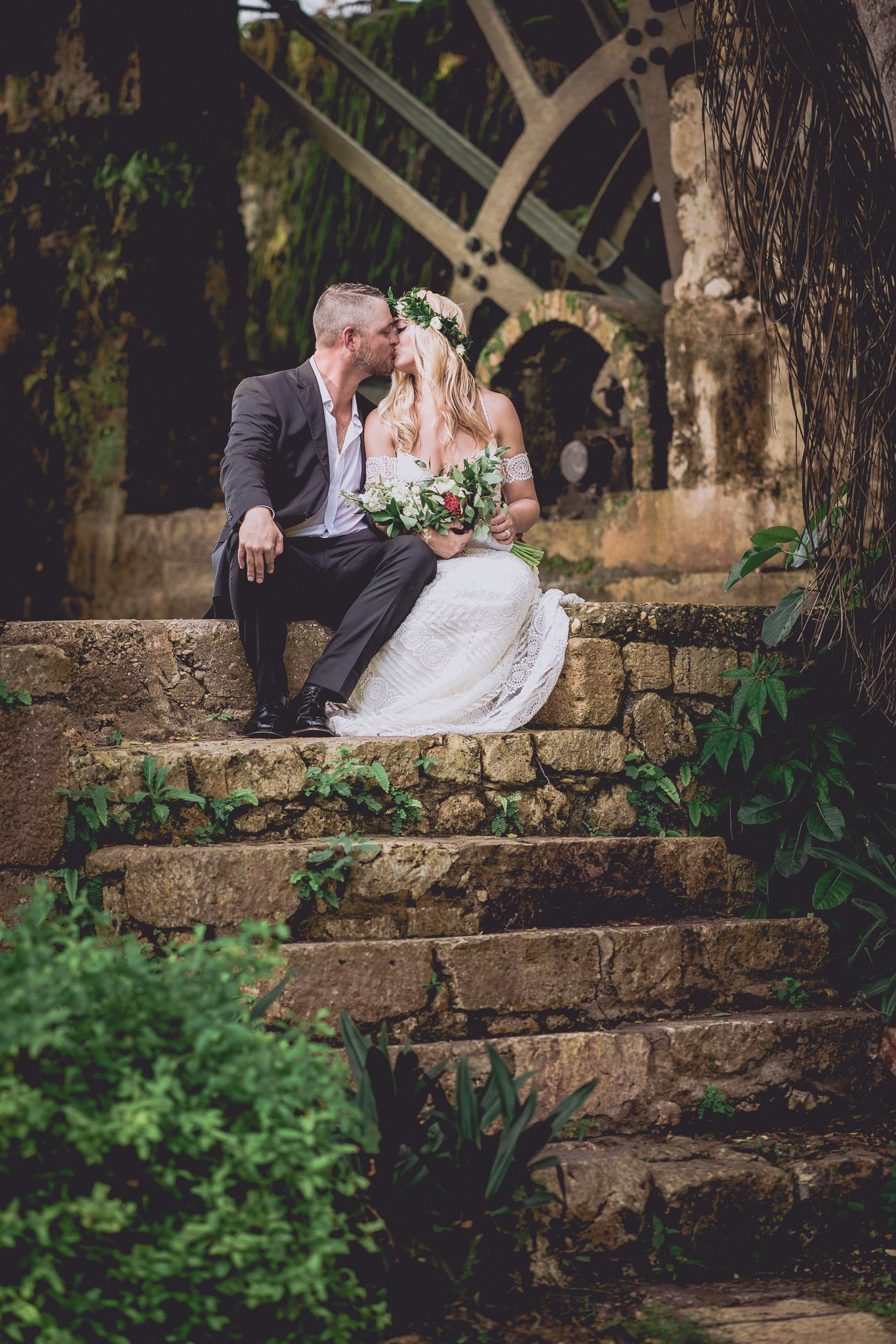 Bride and groom share a romantic kiss on stone steps surrounded by lush greenery in an outdoor garden wedding setting.