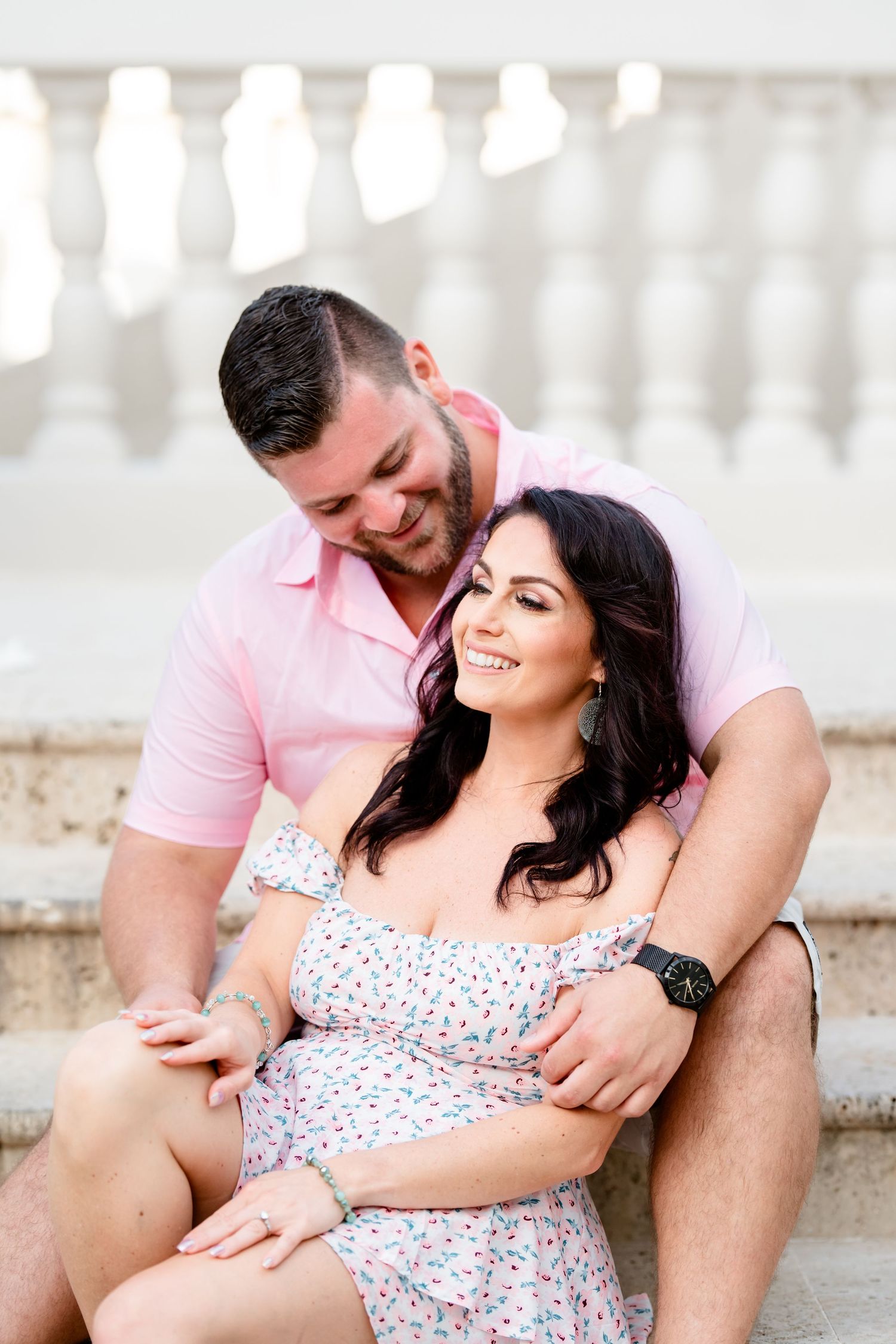 A couple sits together on stone steps outdoors, sharing an intimate moment in matching pink and floral outfits.
