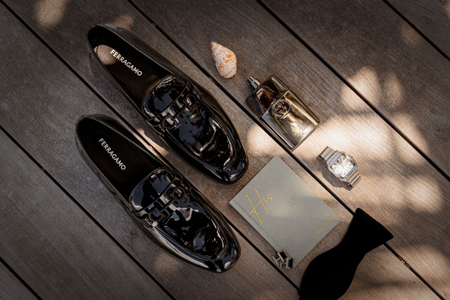 Ferragamo black patent leather loafers displayed with cologne, watch, and accessories on wooden surface.