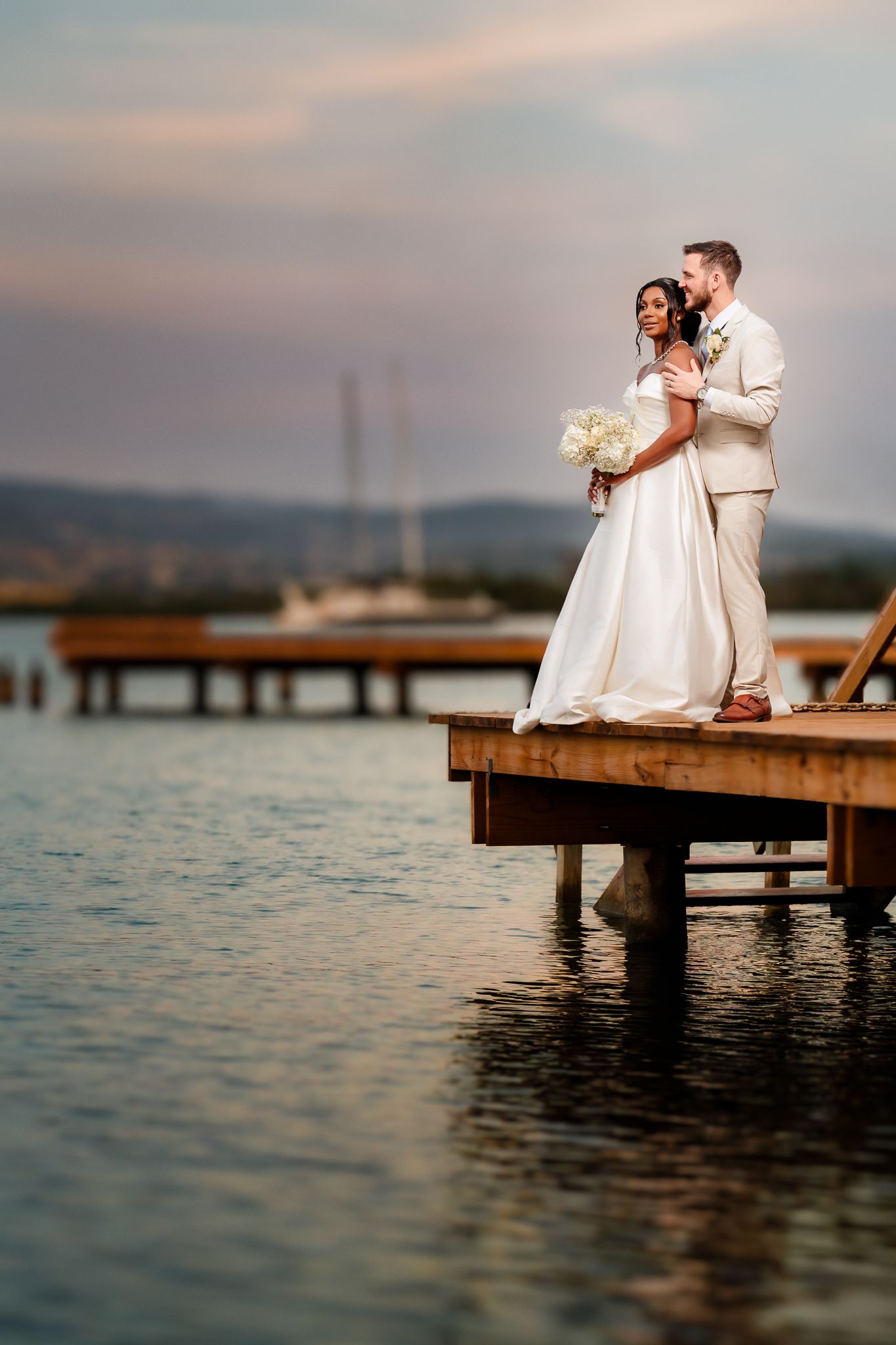 Bride and groom embrace on a wooden dock at sunset, with calm water and hills in the background.