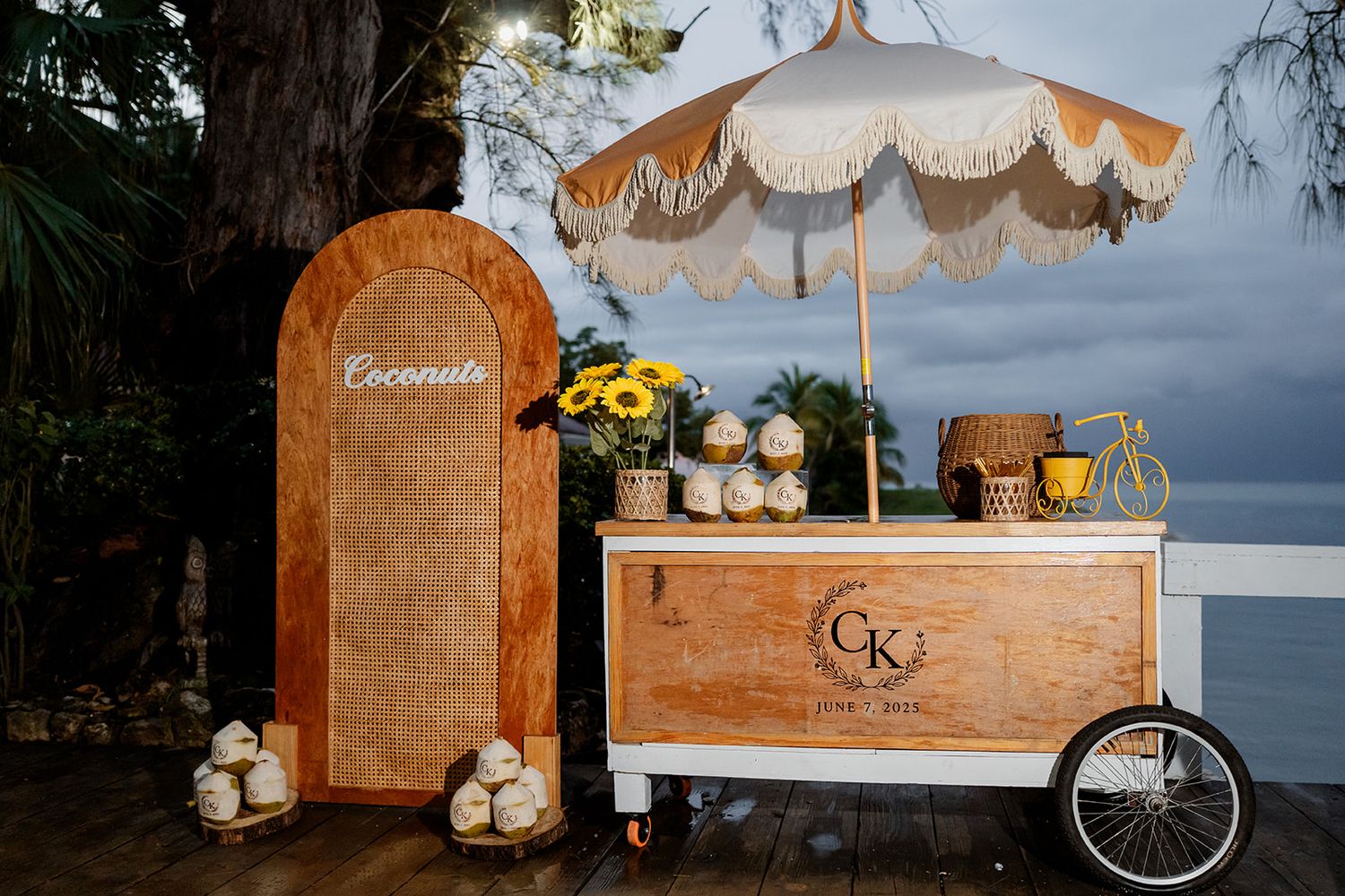 A vintage-style wooden drink cart with parasol umbrella and wicker screen beside it creates a charming beach bar setup.