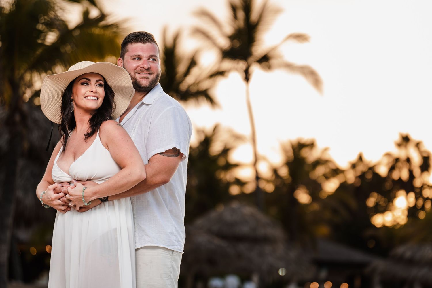 A couple in white clothing embrace during sunset at a tropical beach resort with palm trees silhouetted in the background.