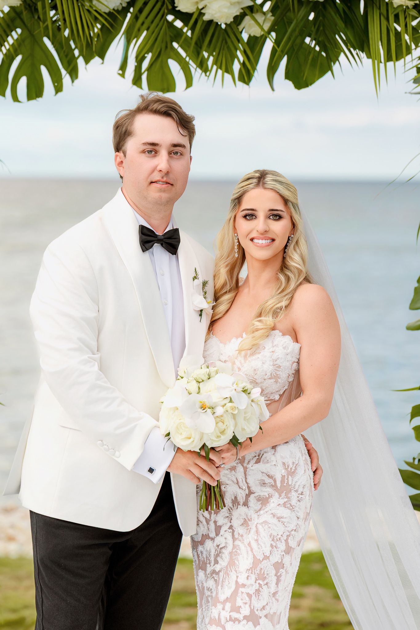Bride and groom pose together outdoors near the ocean, she holds white roses bouquet, he wears white tuxedo jacket.