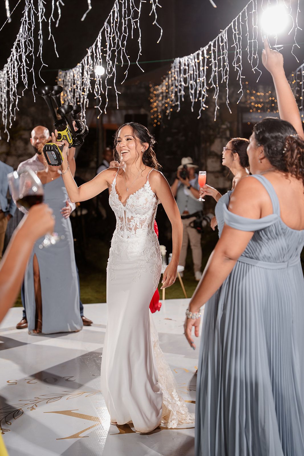 Wedding guests in white and dusty blue dresses dance together on a white dance floor beneath sparkling decorations.