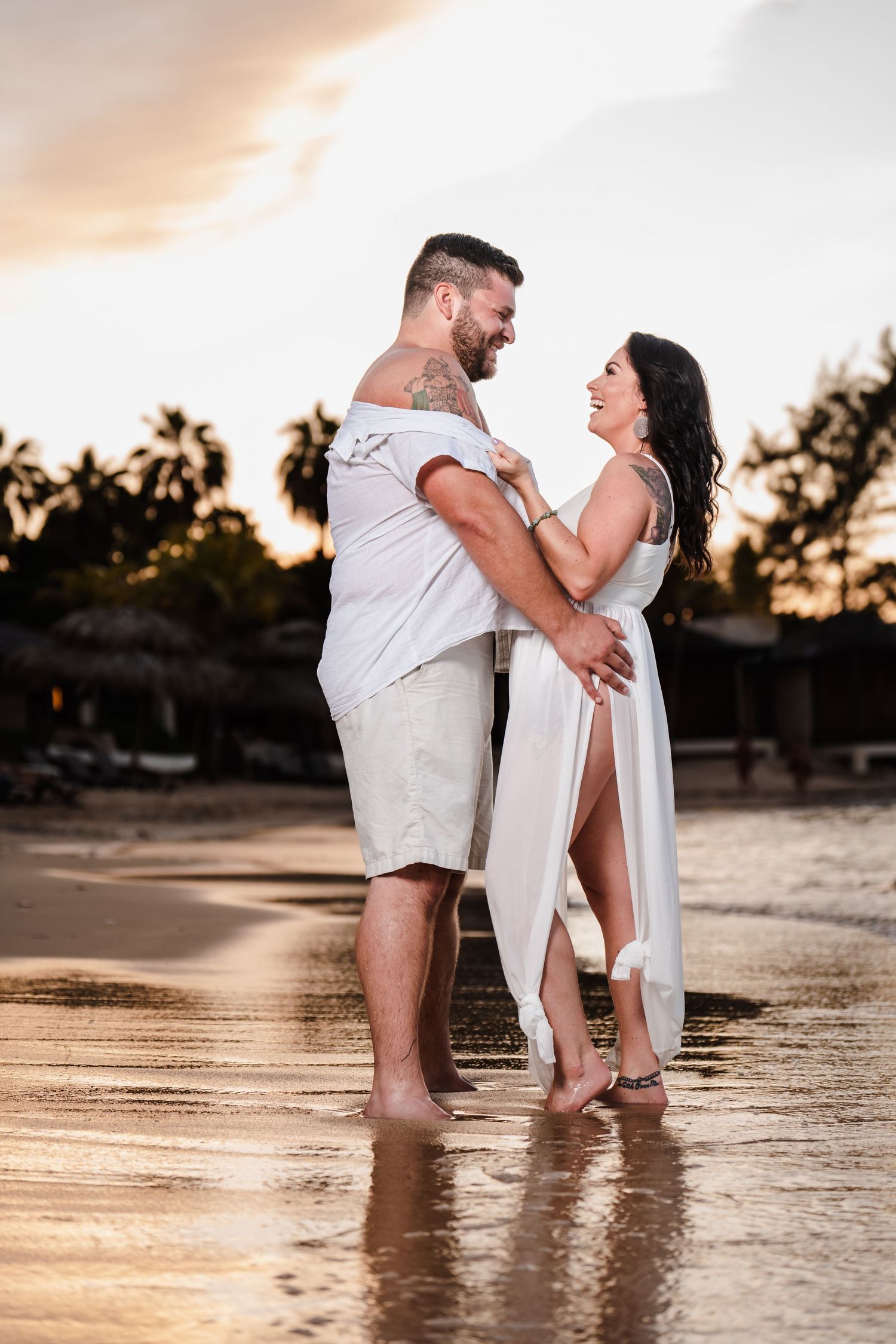 Romantic couple embracing and laughing on a sunset beach with palm trees silhouetted in the background.