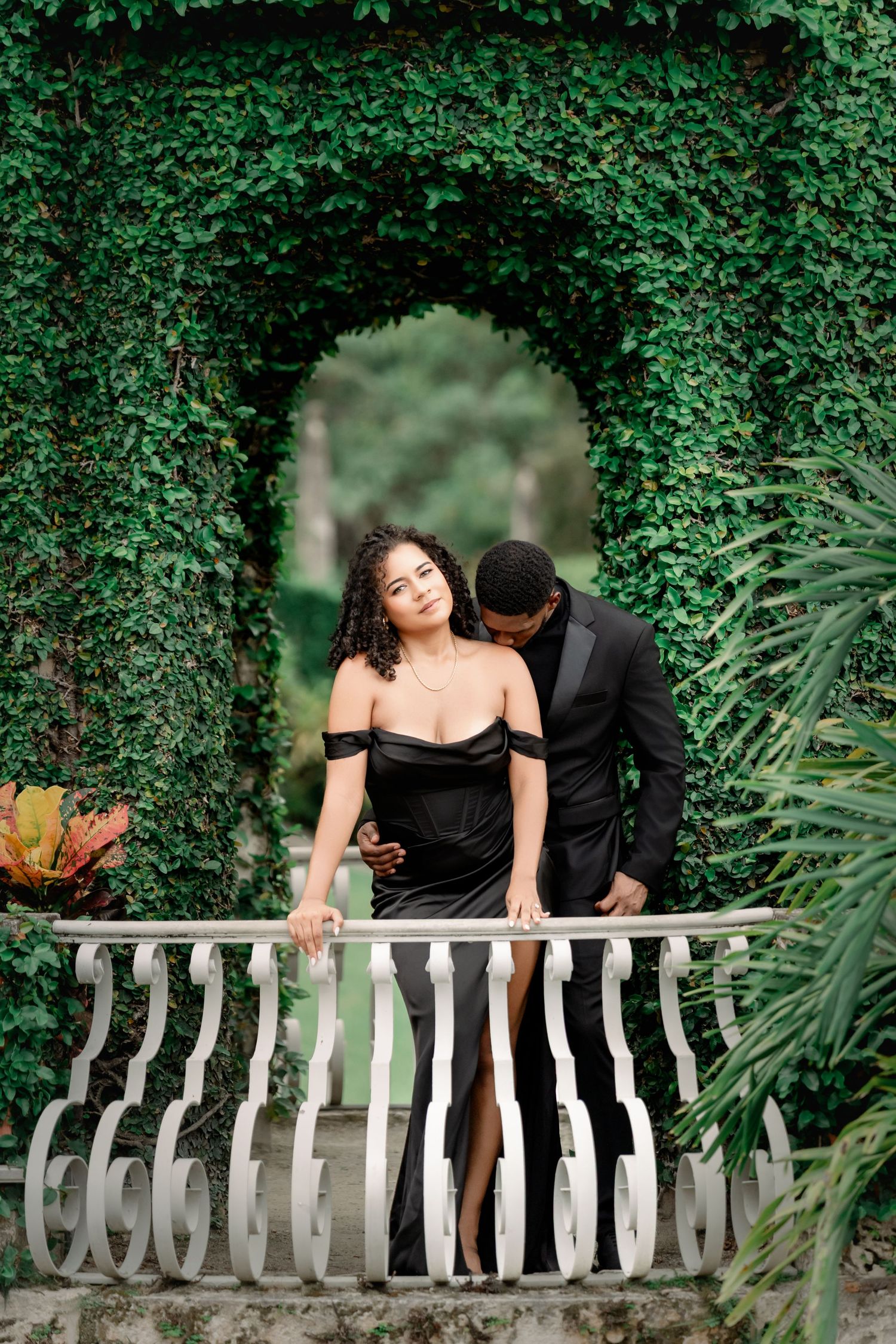 Romantic couple in formal attire share intimate moments on a white stone balcony surrounded by lush green foliage.