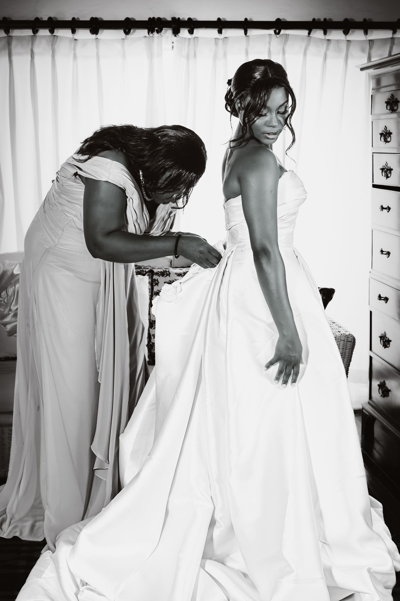 Black and white photo of a bride in a white gown being helped by a bridesmaid in a elegant indoor setting.