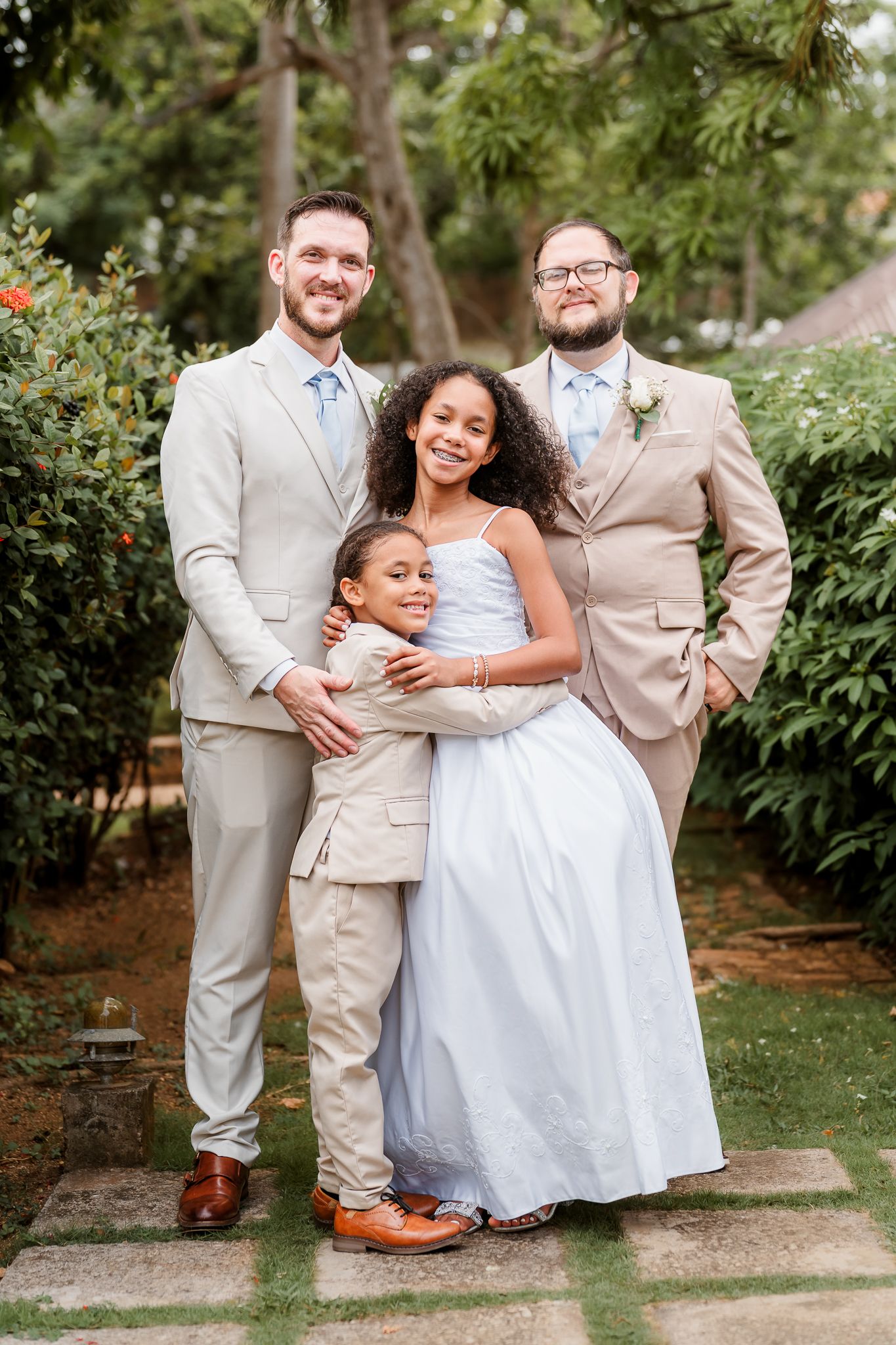 Two grooms in tan suits pose with a girl in a blue dress and young boy at an outdoor wedding ceremony.