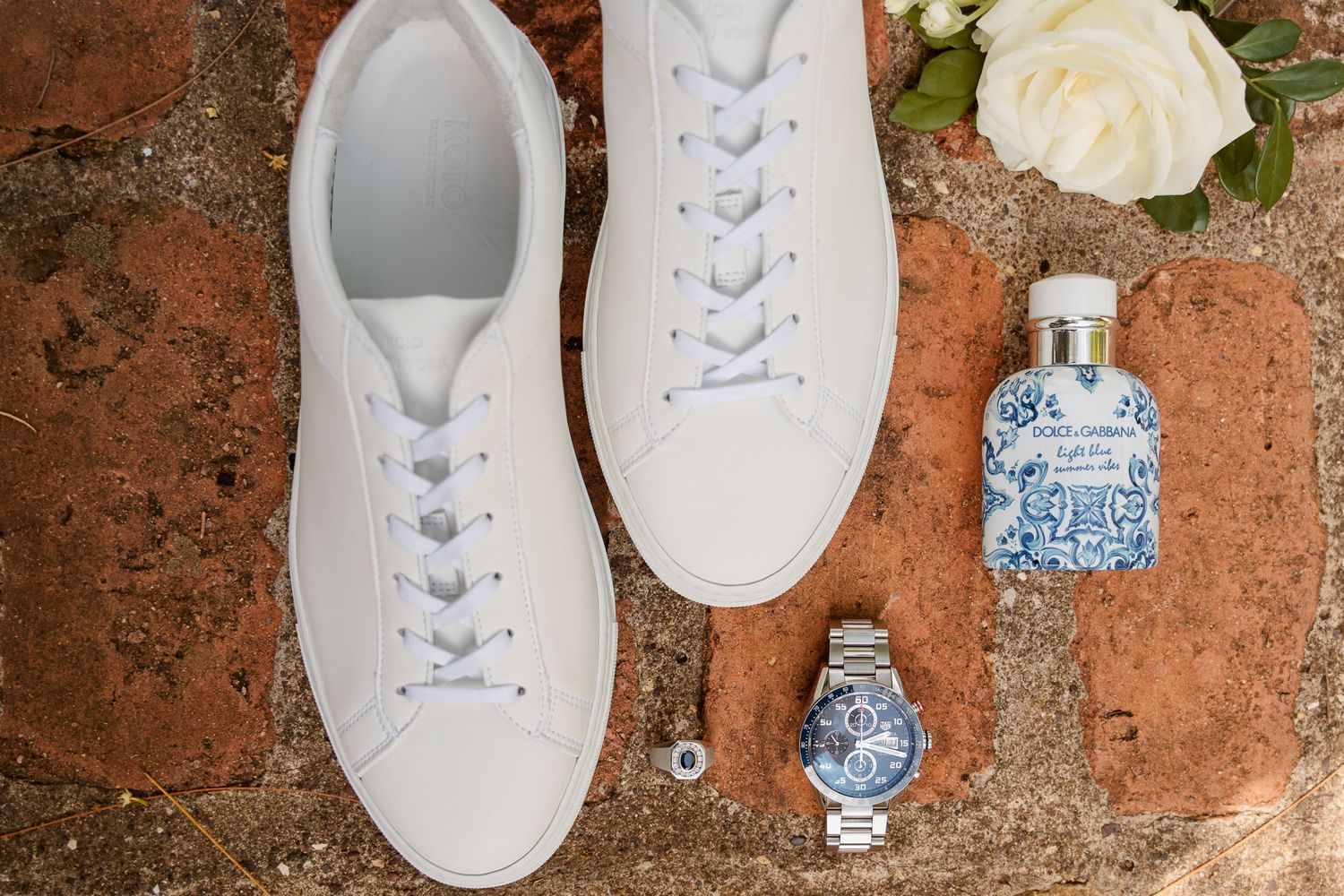 White leather sneakers arranged with a wristwatch, perfume bottle, and cream-colored rose on a rustic terrace surface.